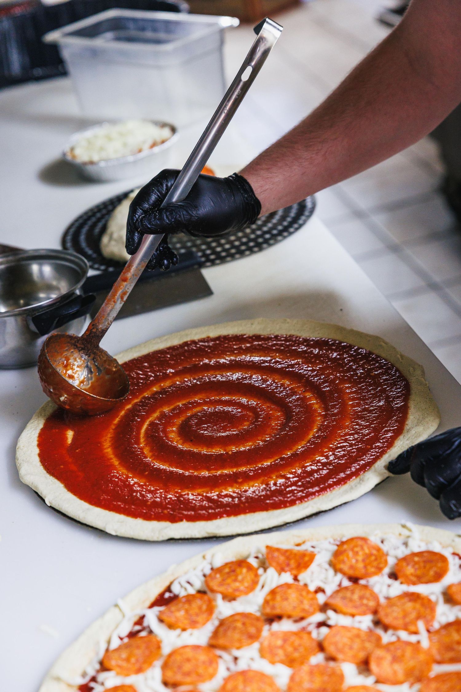 Chef spreading tomato sauce in spiral pattern on pizza dough, with pepperoni pizza visible in foreground.