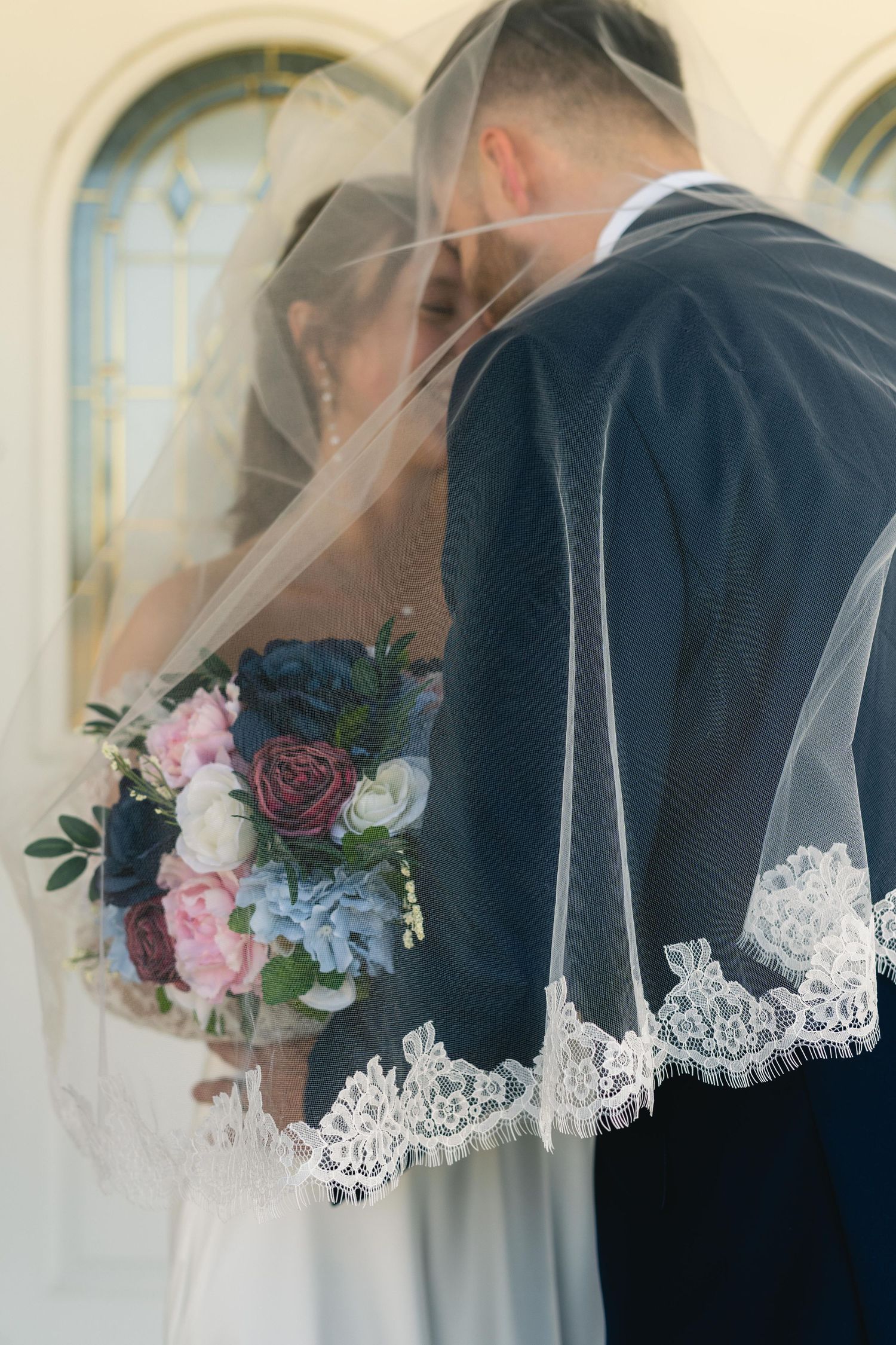 Bride and groom sharing a romantic kiss under a lace-trimmed veil, bride holding a bouquet of pink, blue, and burgundy flowers.