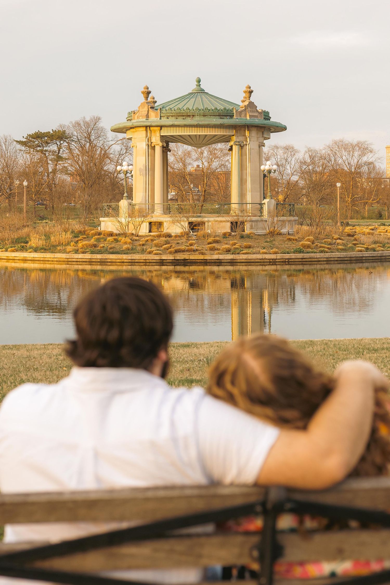 Couple sitting on a bench admiring a classic gazebo reflected in a calm pond in a serene autumn park.