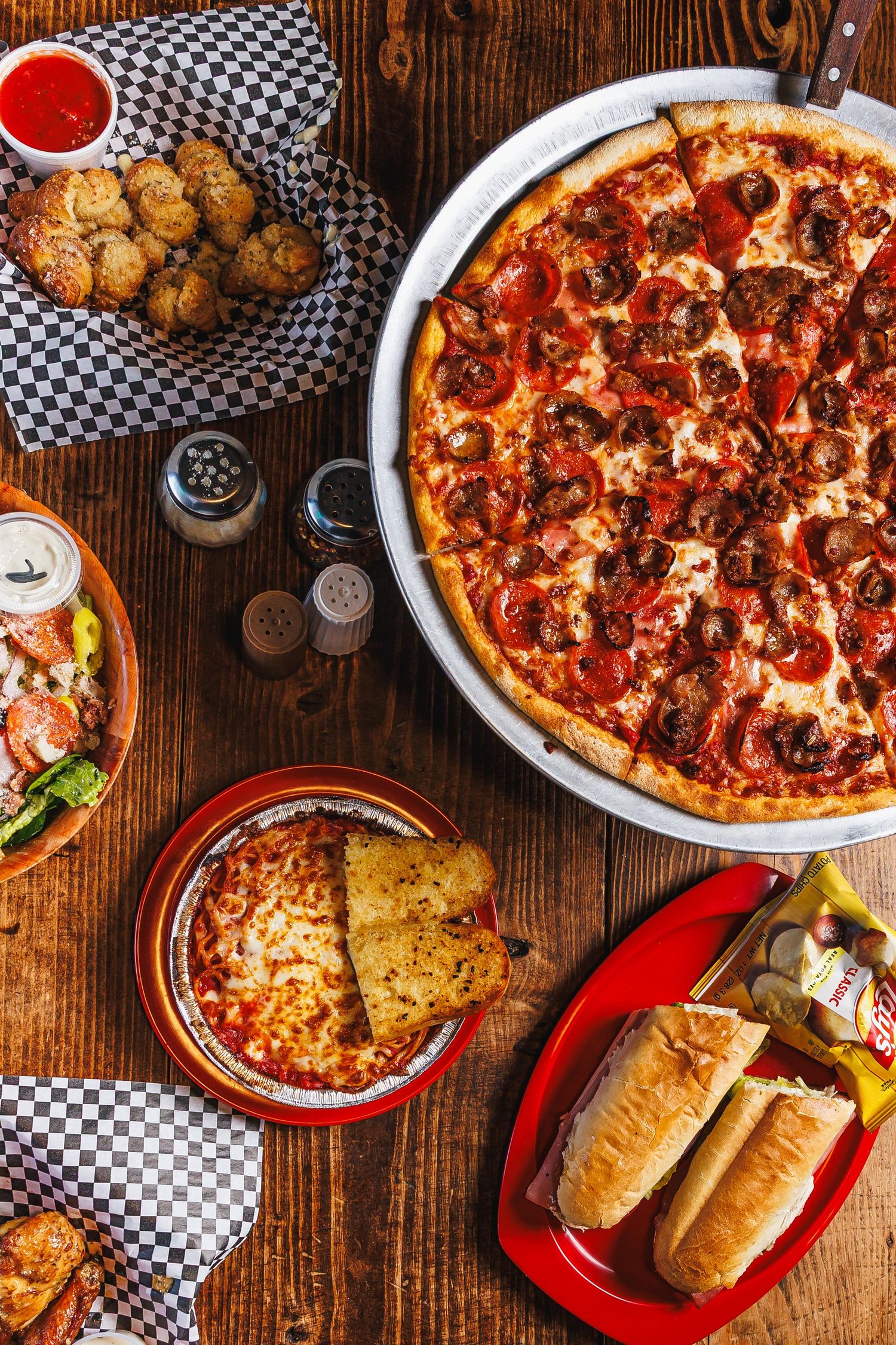 Overhead view of pepperoni pizza, sandwiches, breadsticks and appetizers on a rustic wooden table at an Italian restaurant.