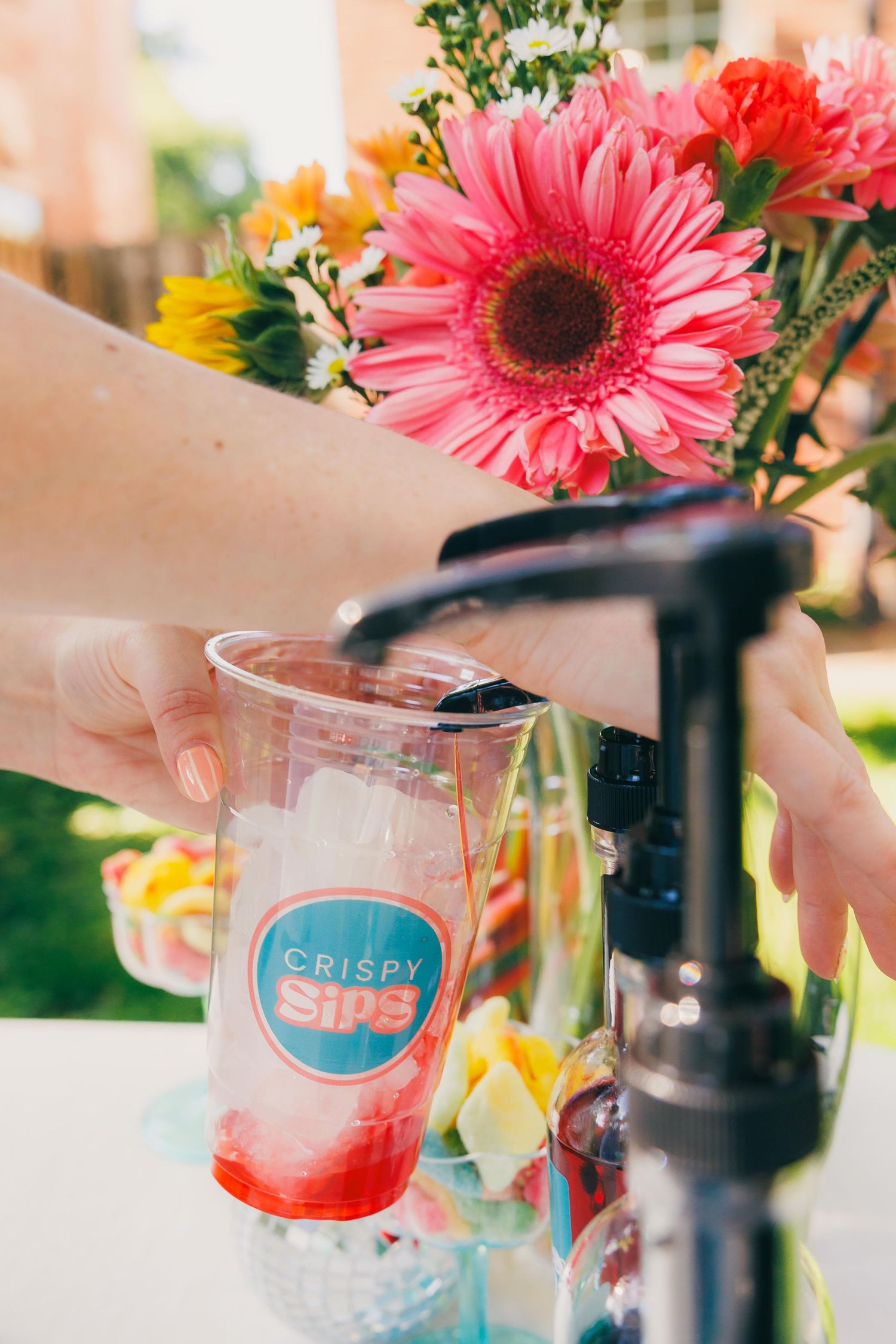 Hand filling a Crispy Sips branded cup with ice at an outdoor event, with vibrant pink gerbera daisies in the background.