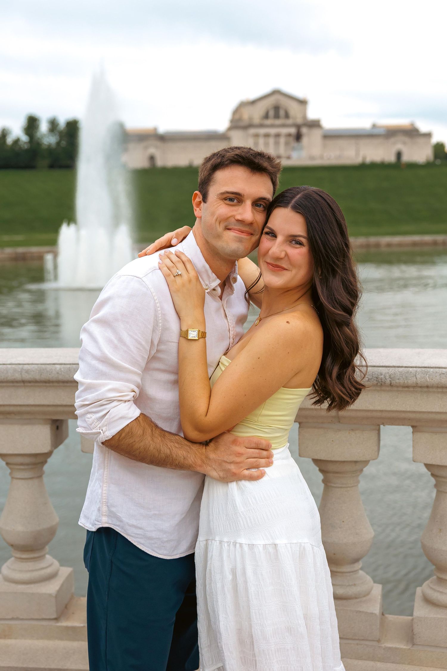 Couple embracing and smiling in front of a fountain and classical building outdoors.