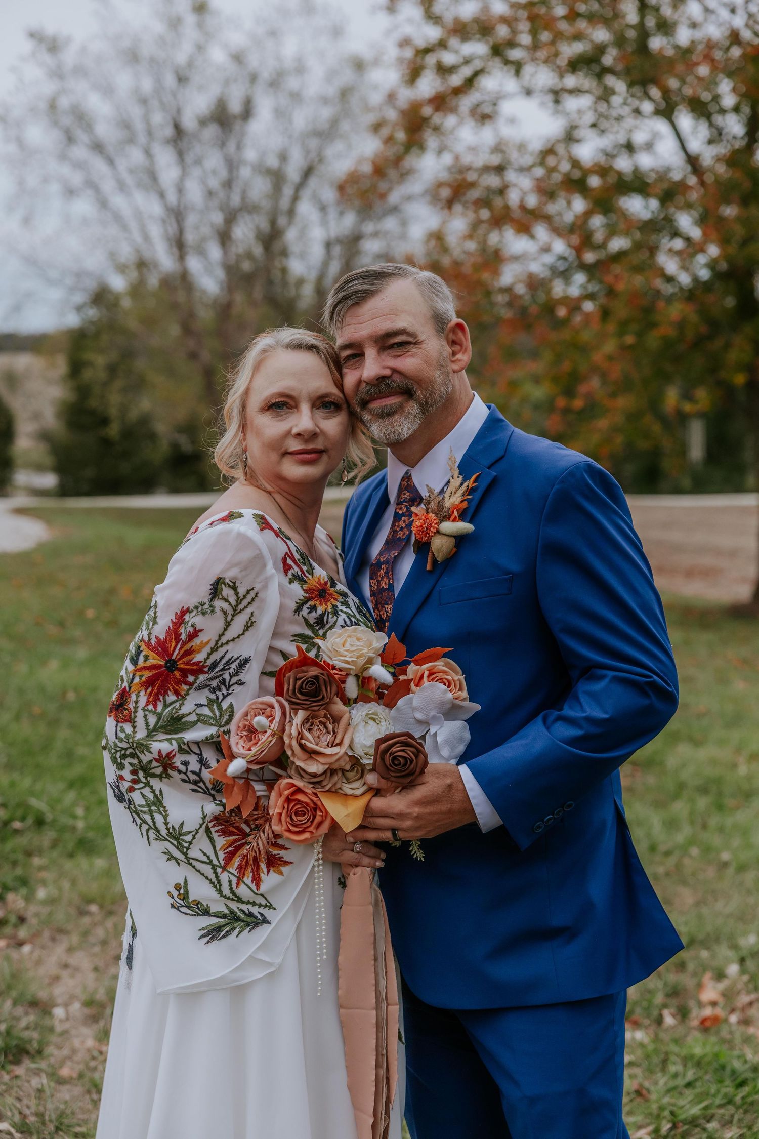Wedding couple poses outdoors with autumn foliage, featuring a white dress with floral details and navy suit in Missouri.