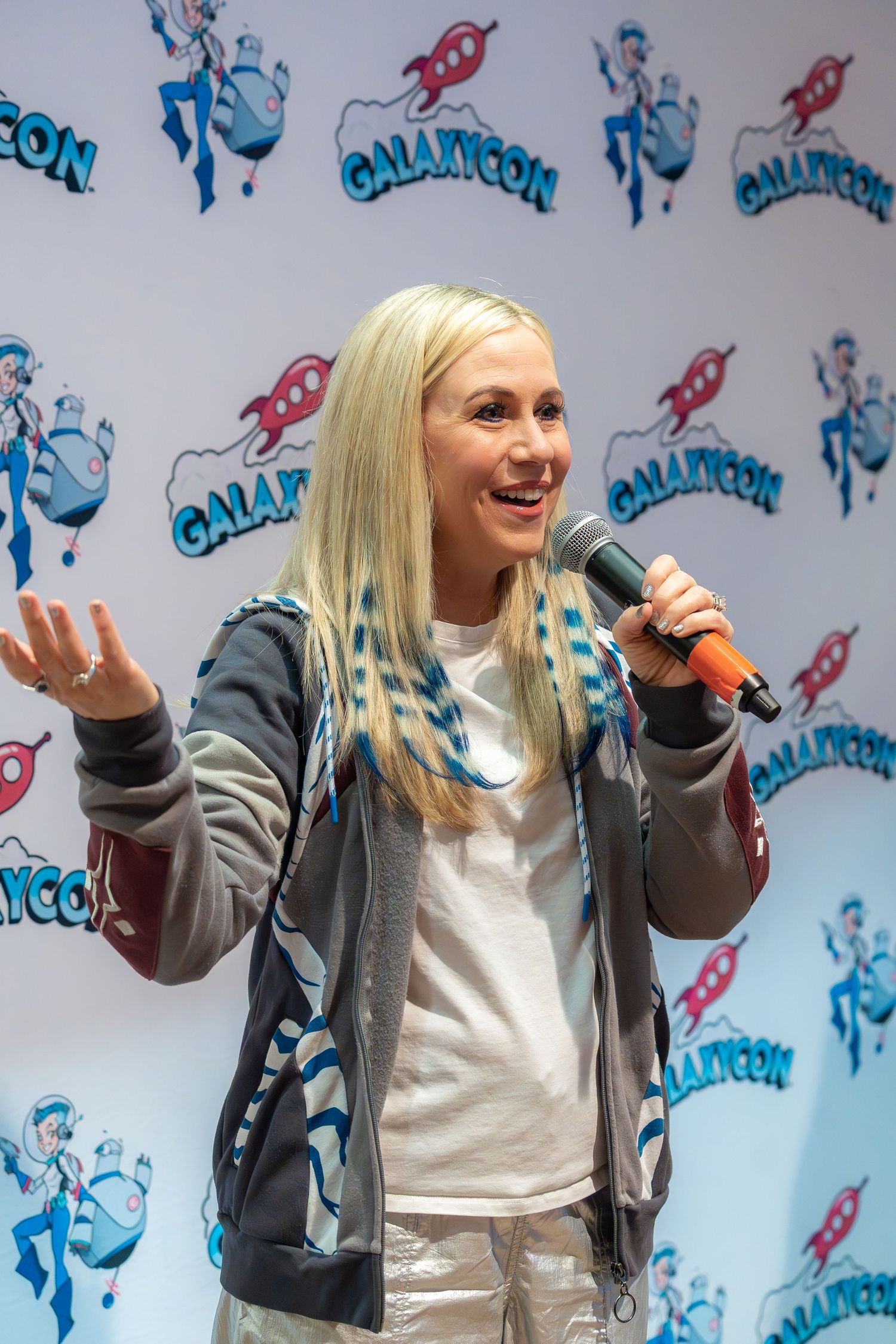 Ashley Eckstein in gray hoodie and white shirt speaking into microphone against blue and white patterned backdrop.