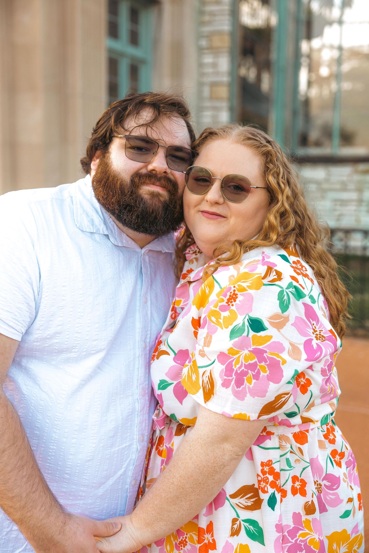 Couple in sunglasses posing outdoors, man in white shirt with beard, woman in colorful floral dress, smiling together.