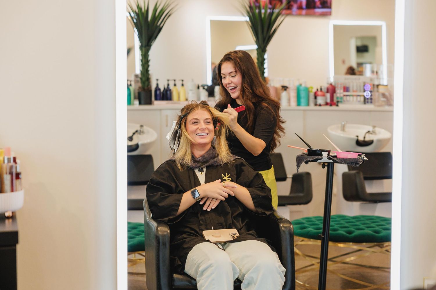 Smiling woman getting hair styled by brunette hairdresser in modern bright salon with mirror lights.