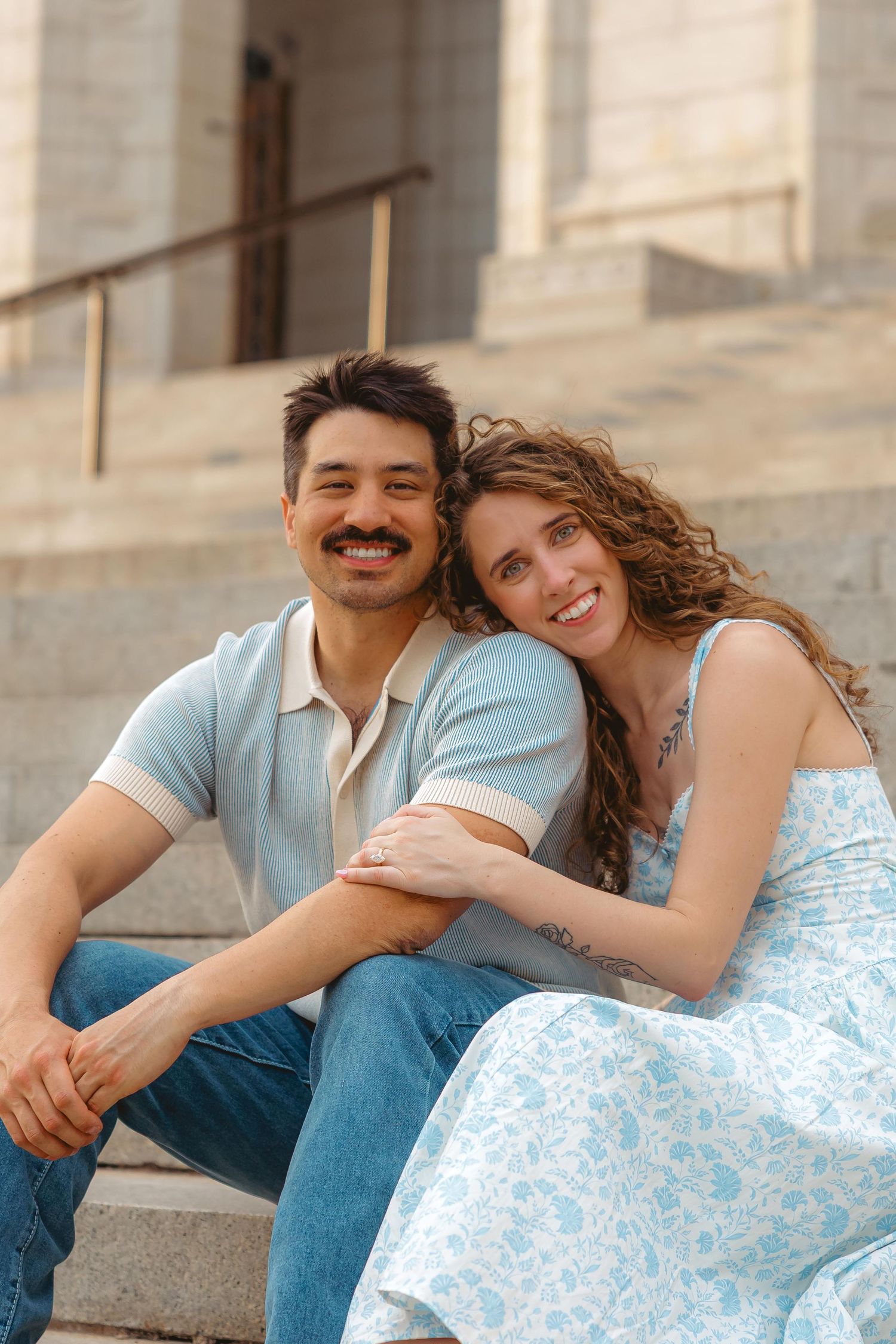 Happy couple sitting on stone steps, woman in floral dress leaning on man in blue polo shirt, smiling outdoors.