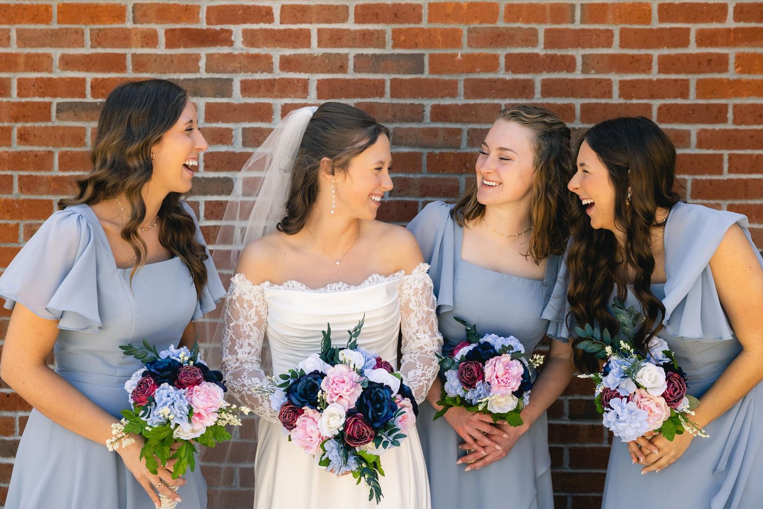 Bride in white lace gown with veil laughs with three bridesmaids in blue dresses holding pink and navy bouquets.