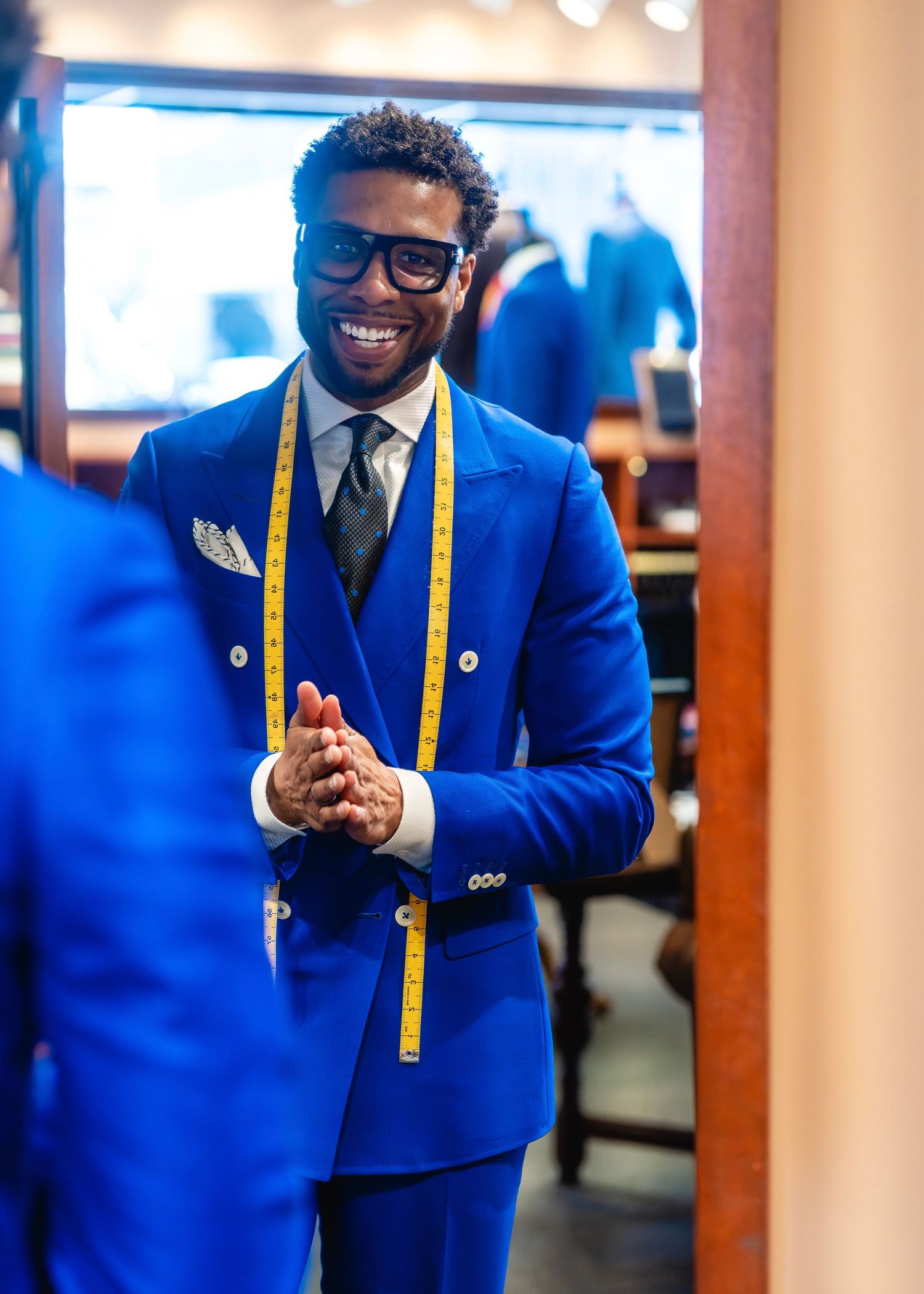 A tailor in a bright blue suit with measuring tape smiles in a clothing store in Clayton, Missouri.