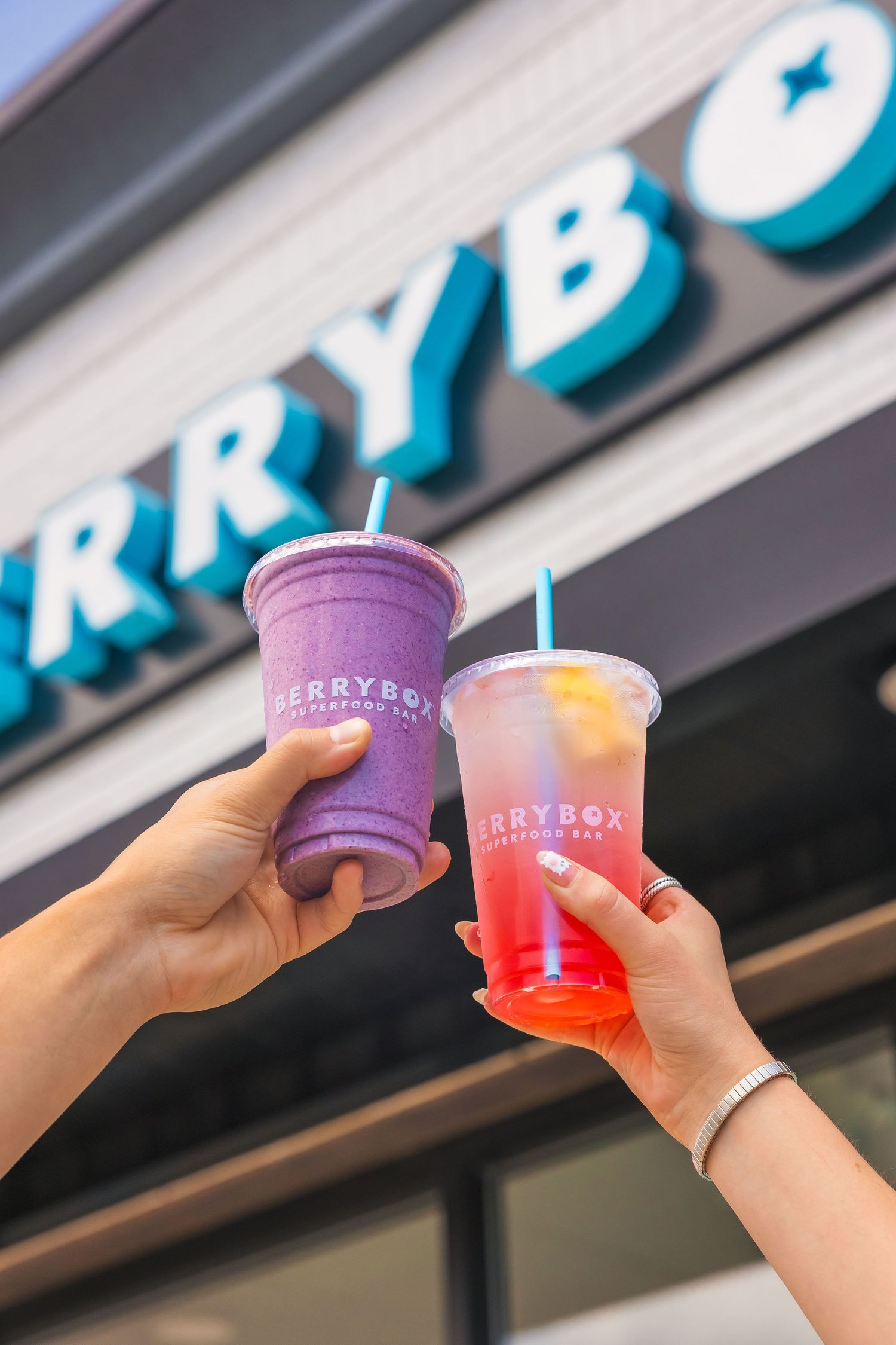 Two hands holding colorful BerryBox smoothie drinks outside a BerryBox juice bar location.