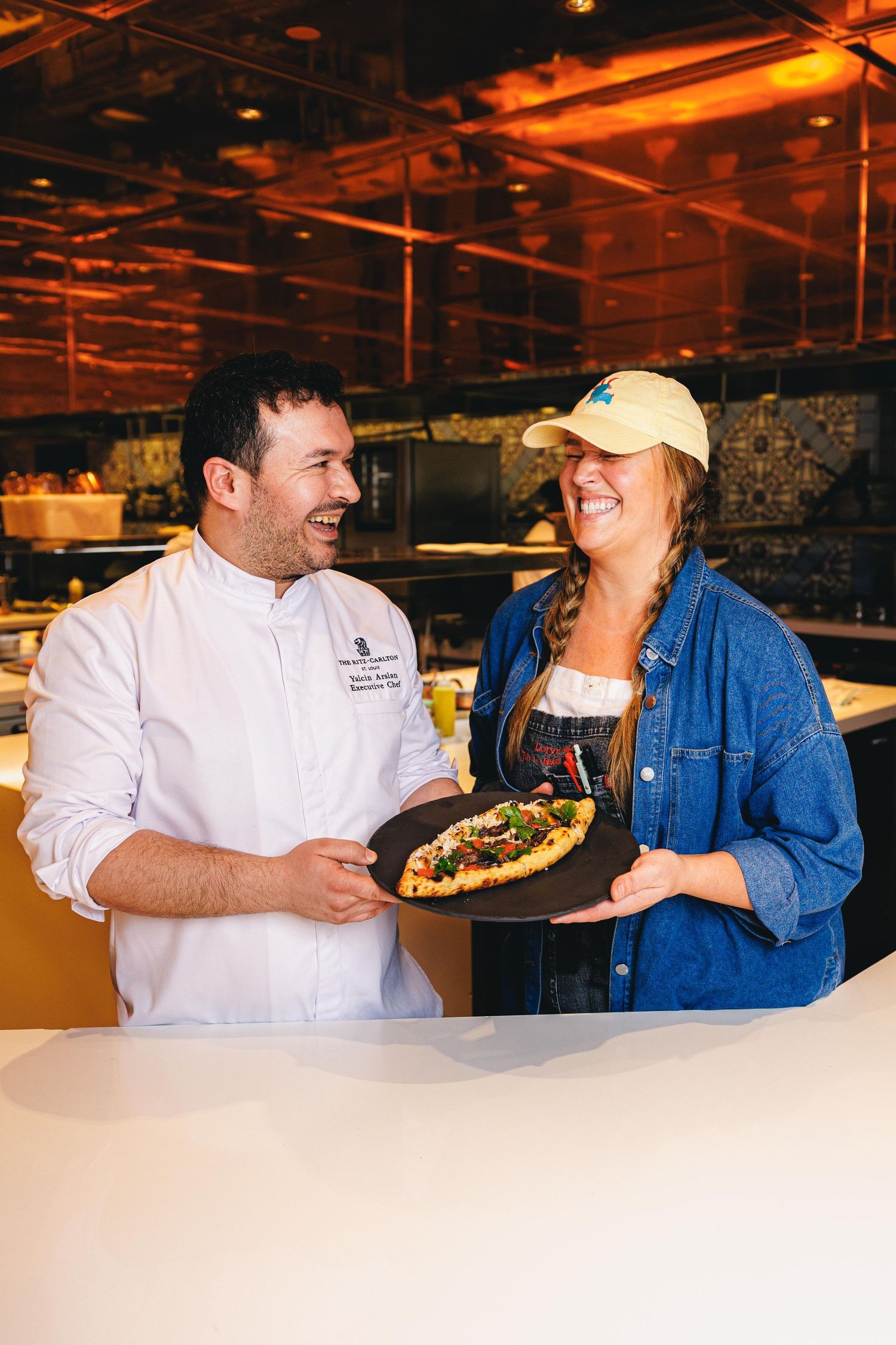 A chef in white coat and casual diner share a laugh while presenting a gourmet dish in a warmly lit restaurant.