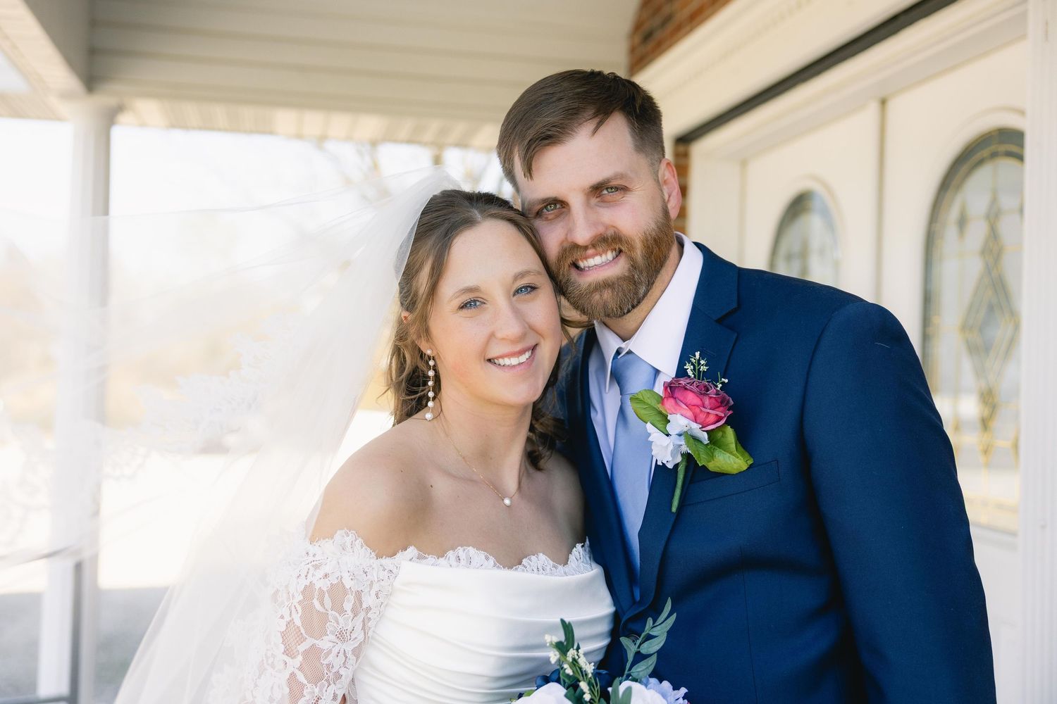 Happy bride and groom posing together on wedding day, bride in lace off-shoulder gown, groom in navy suit with floral boutonniere.