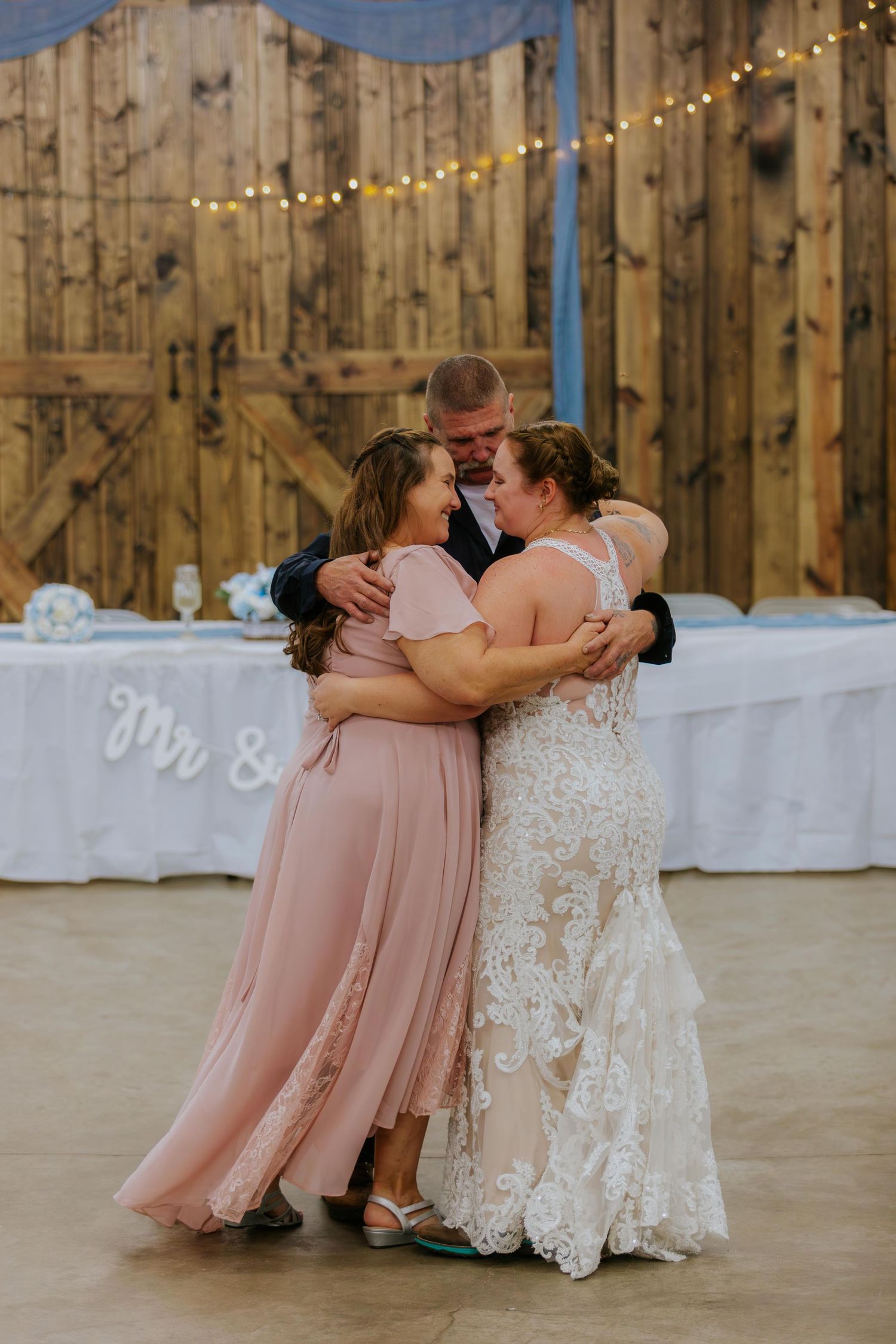 Wedding party in pink bridesmaid dresses and white lace bridal gown pose together in rustic barn venue with string lights.