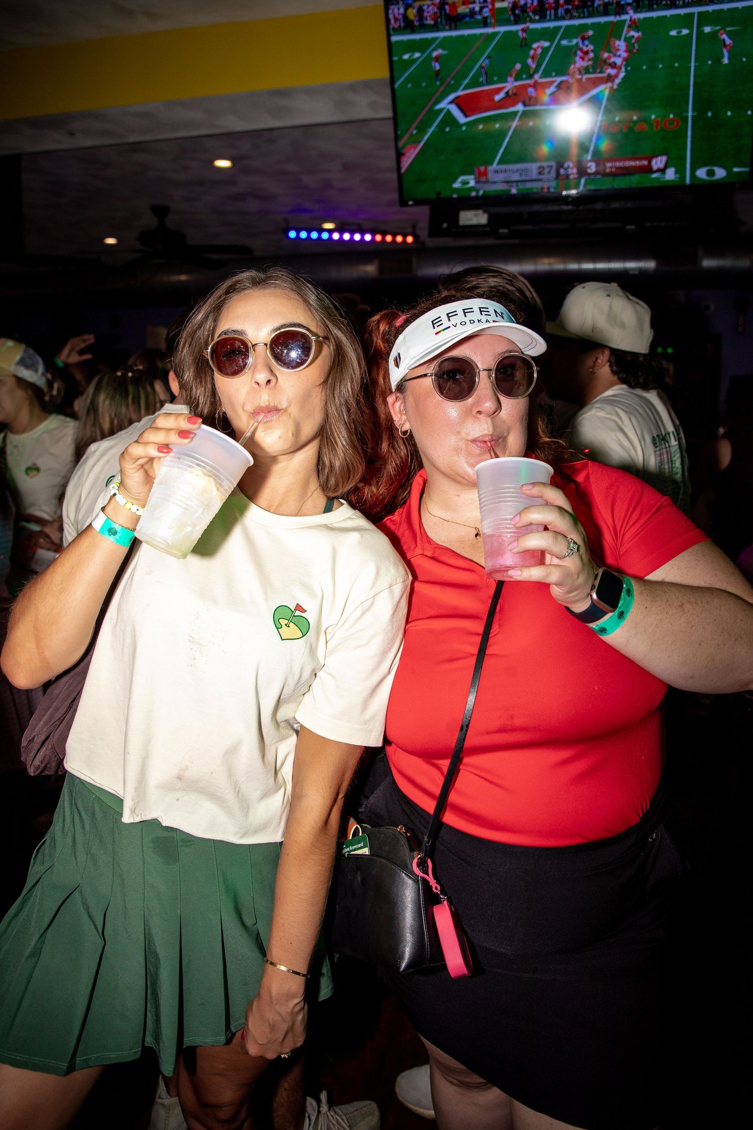 Group wearing green skirts and sunglasses drinking at a bar with TVs showing sports.