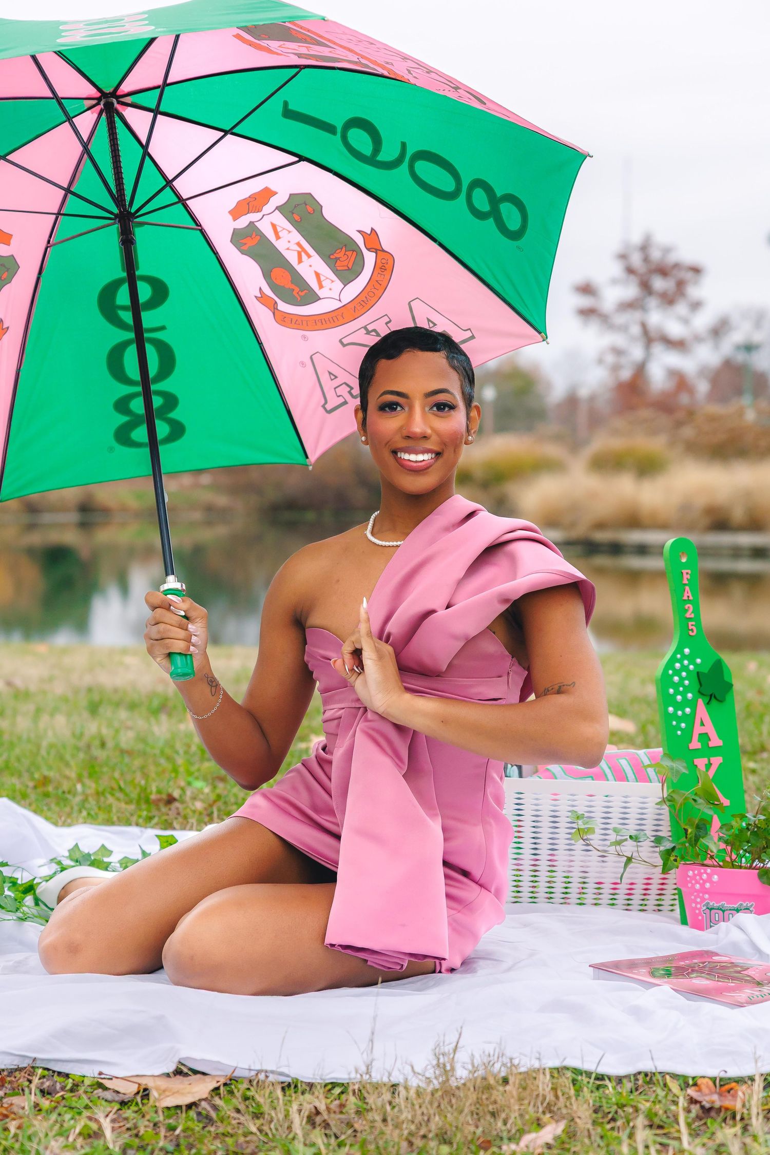 Pink and green sorority themed picnic setup with umbrella and blanket on grass near a lake.