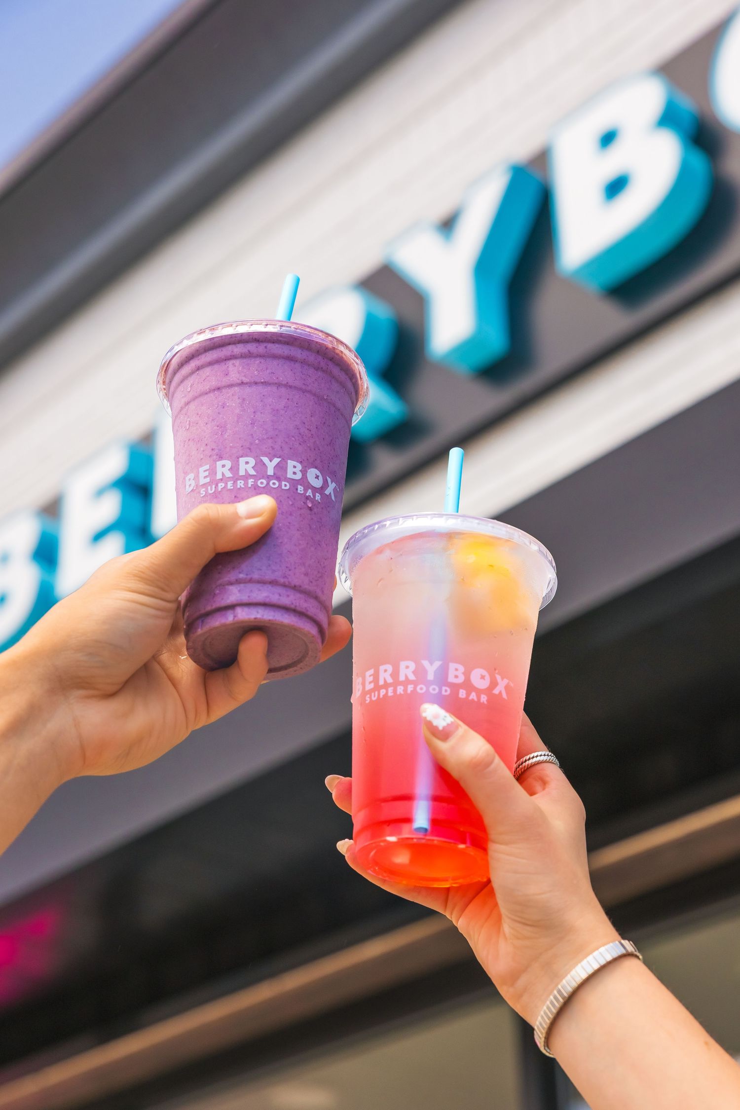 Two hands holding BerryBox Superfood Bar drinks, a purple smoothie and a pink lemonade, in front of the colorful store sign.