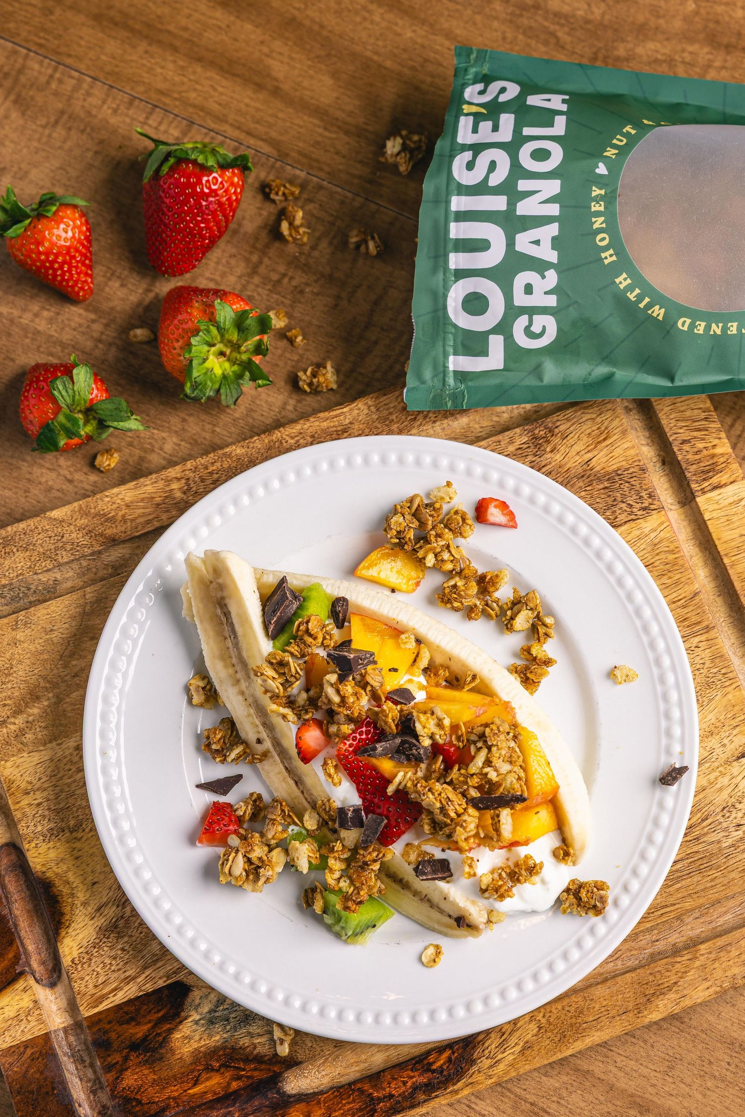 Banana split with granola, strawberries, and fruit on white plate beside Louise's Granola bag on wooden board.