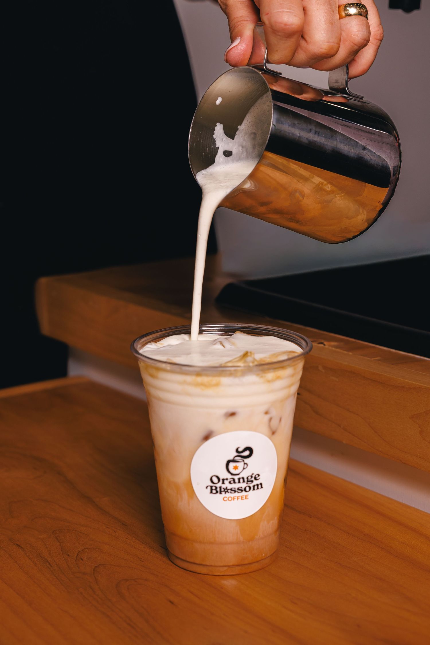 Barista pouring steamed milk into an iced latte in an Orange Blossom Coffee branded cup on a wooden counter.