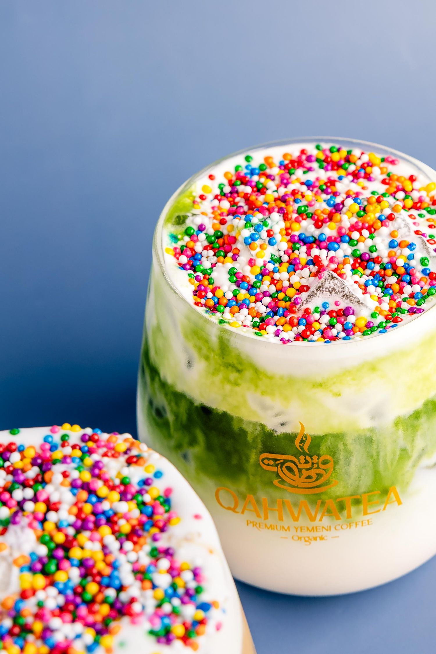 Colorful rainbow sprinkle-topped matcha latte in a glass cup beside a frosted donut, served at Qahwa Premium Coffee.