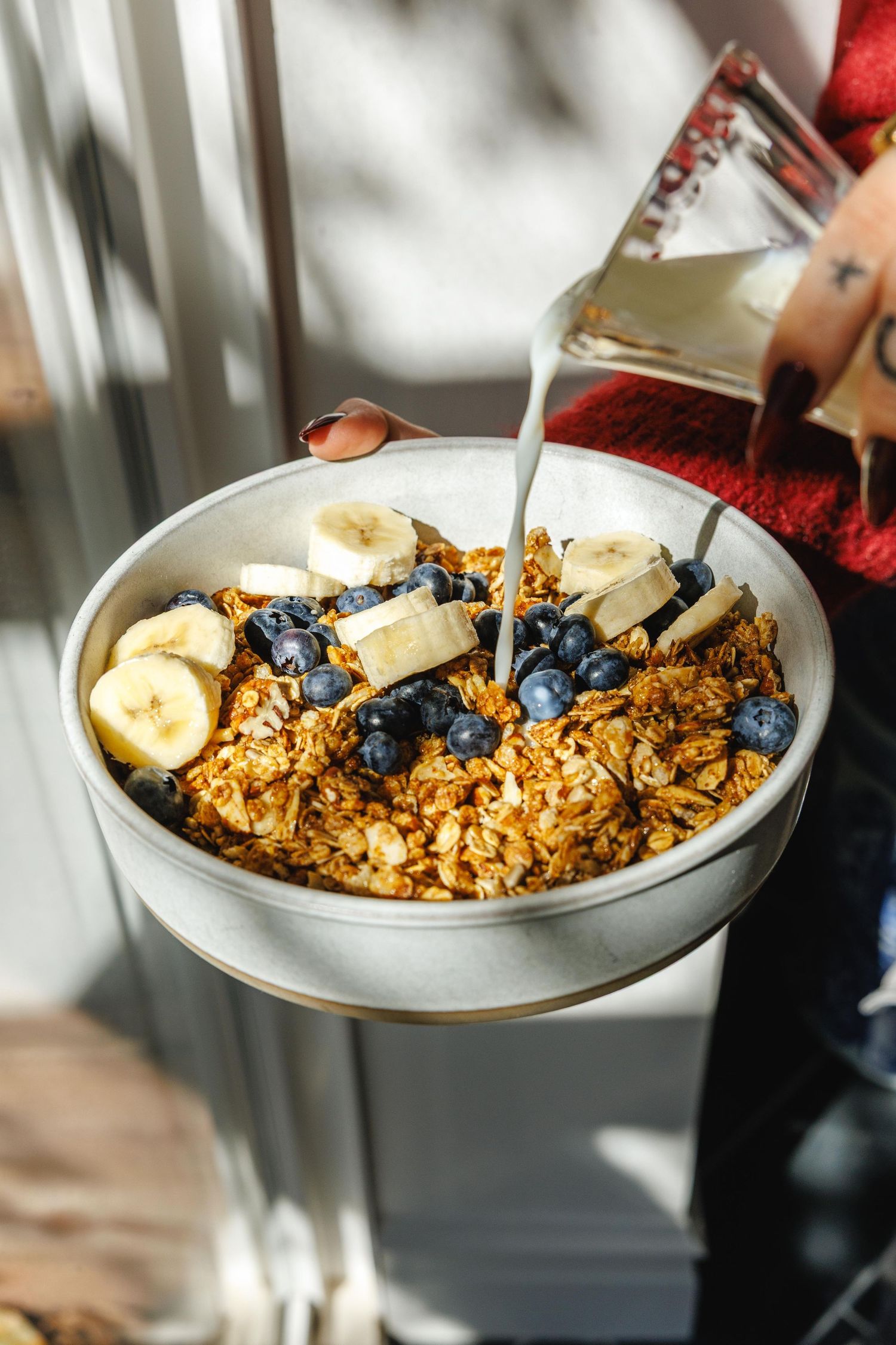 A bowl of granola topped with fresh blueberries and banana slices, with milk being poured over it.