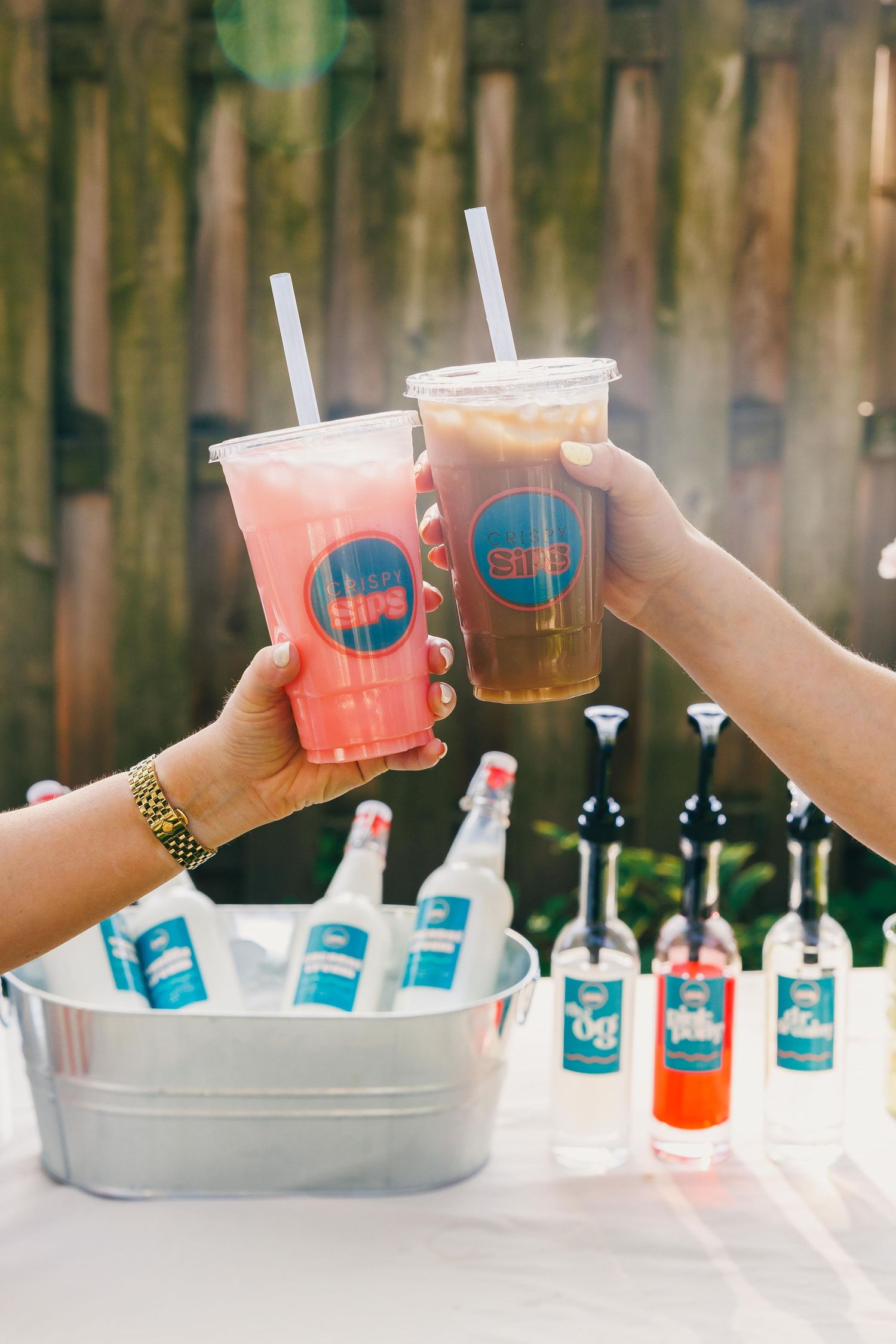 Two hands toasting with branded iced drinks outdoors, with bottles on ice in a metal bucket on a white table.