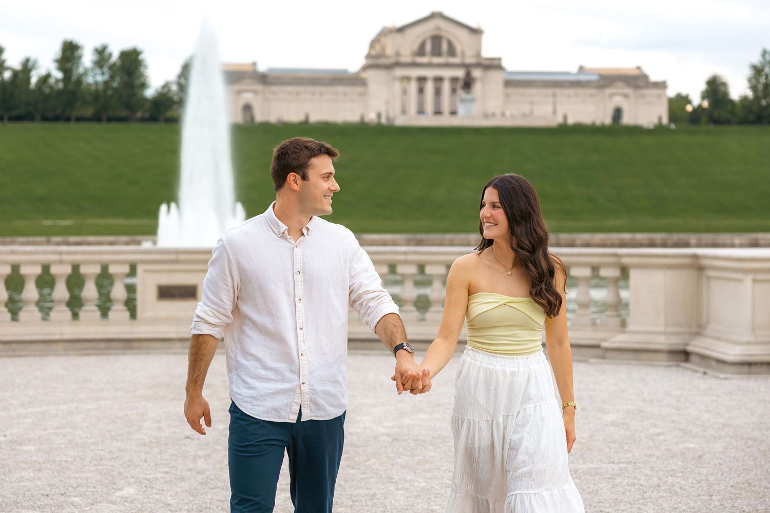 Couple holding hands walking in front of a grand fountain and neoclassical building in a lush green park.