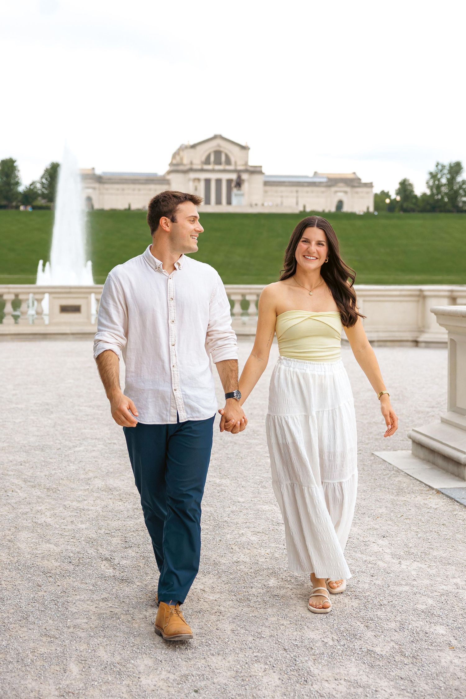 A smiling couple holds hands while walking near a fountain and grand building outdoors.