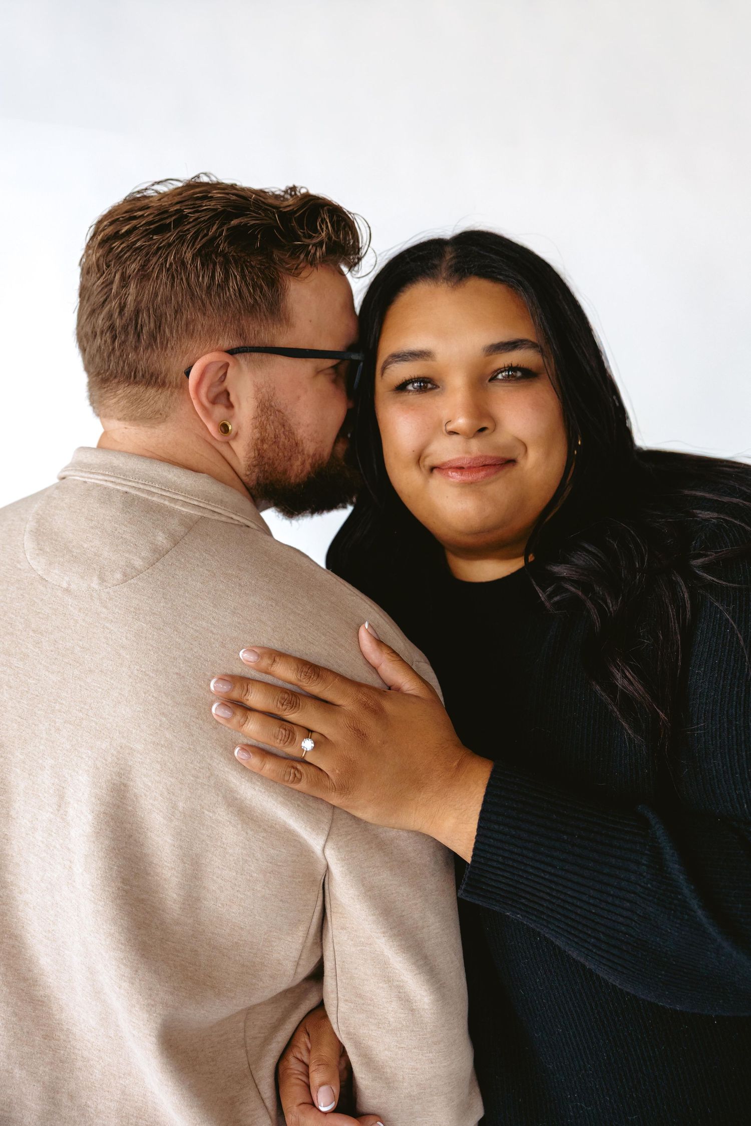 Couple embracing, woman smiling at camera wearing engagement ring, man kissing her cheek, white background.