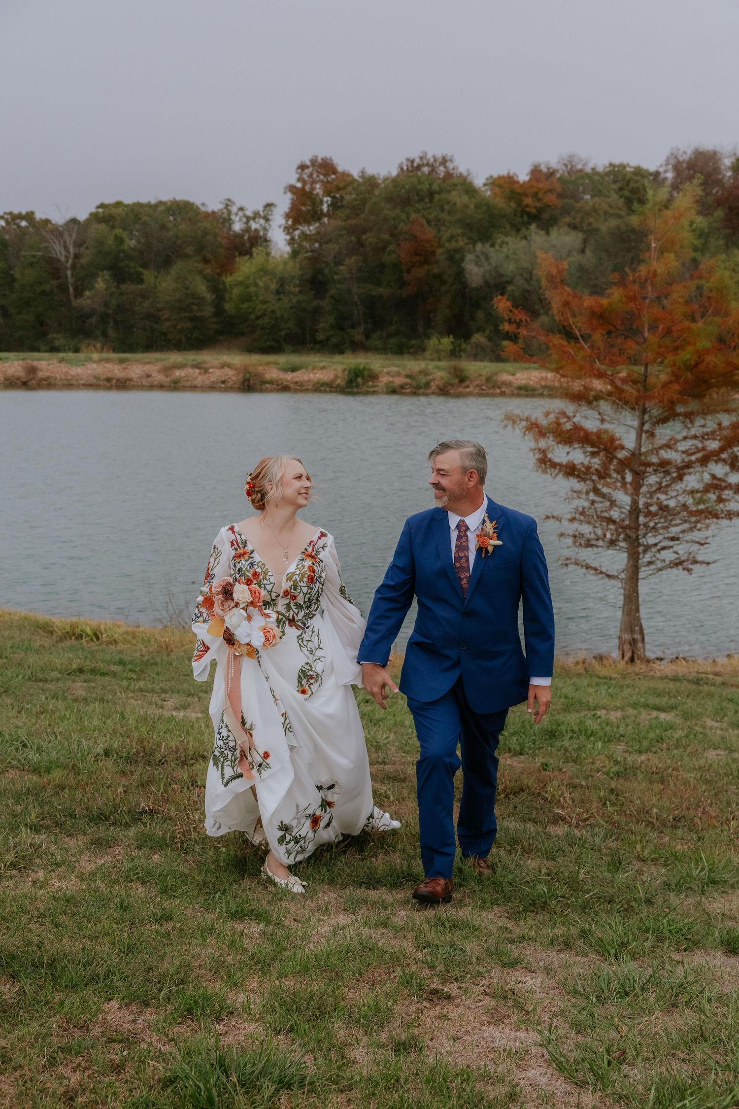 Bride in embroidered dress and groom in blue suit dance by lakeside during fall wedding.