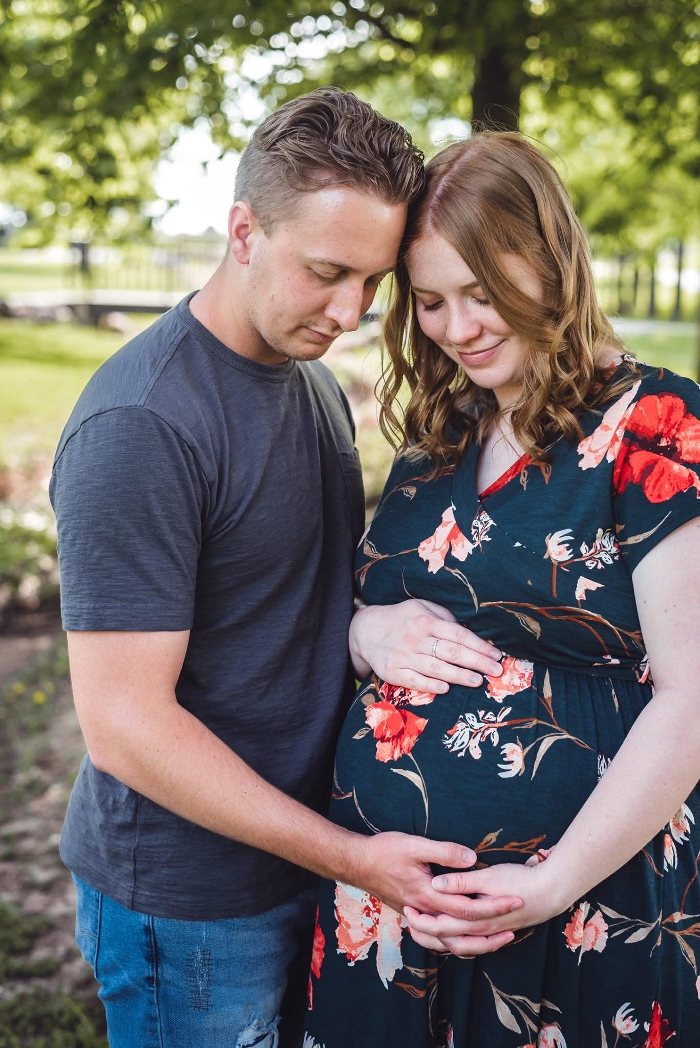 Maternity photo session in a park with couple wearing navy and floral dress touching pregnant belly.