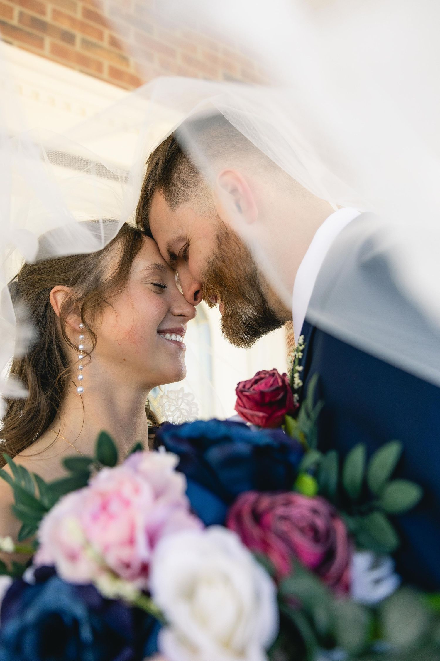 Bride and groom touching foreheads under veil with pink and navy bouquet in foreground.