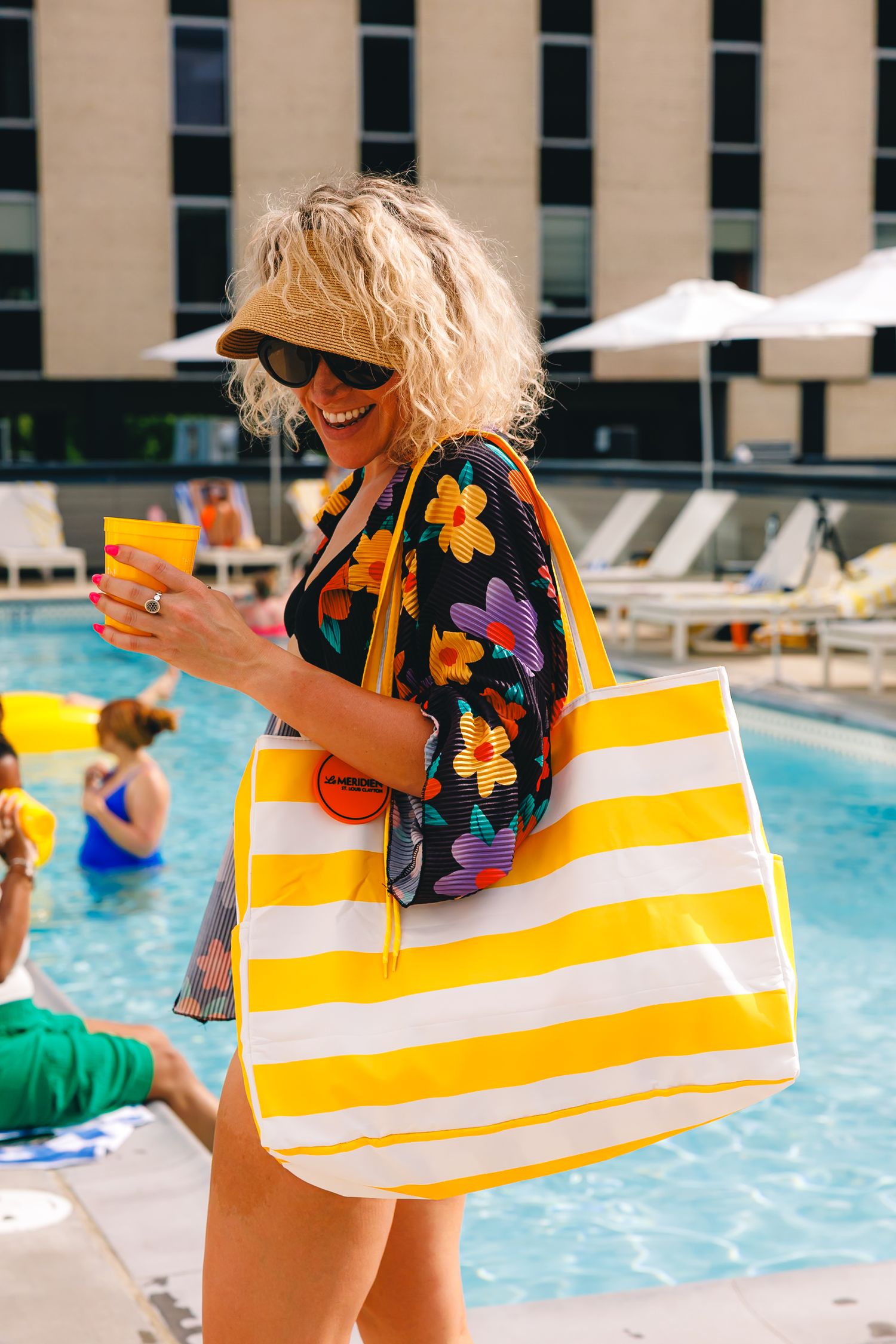 Smiling woman in floral top holding orange drink with yellow striped beach bag by a sunny hotel pool.