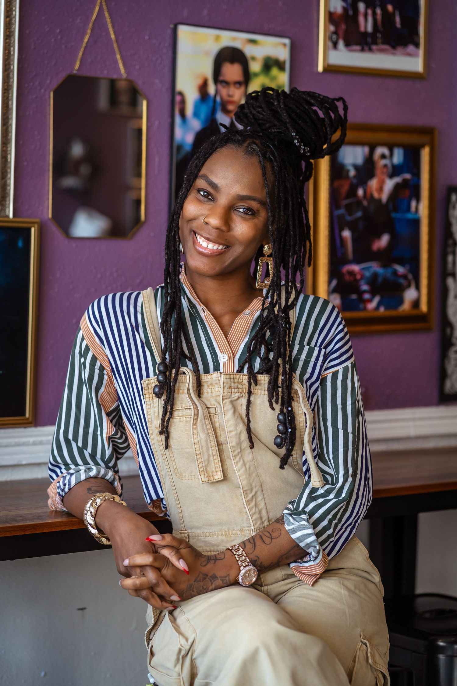 A casual indoor portrait featuring striped shirt and overalls against a purple wall with hanging decor in University City, Missouri.