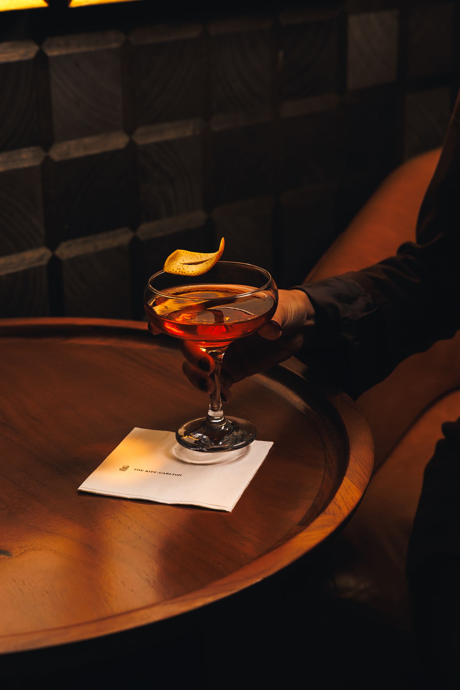Hand holding a red cocktail in a coupe glass with citrus twist garnish on a wooden table in a dimly lit bar.