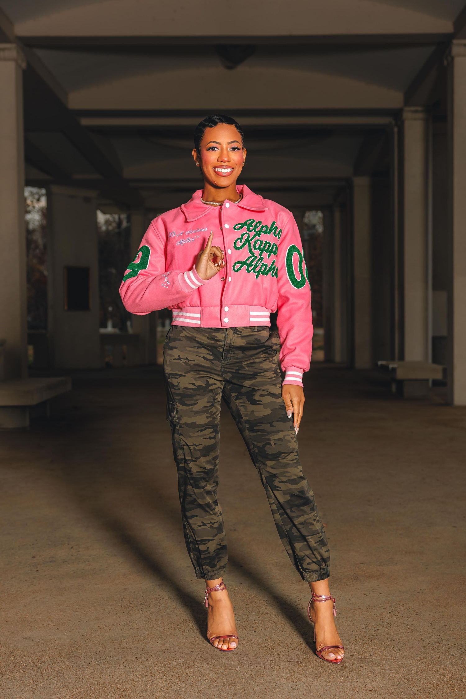 Pink bomber jacket styled with camouflage pants and heels in urban setting.