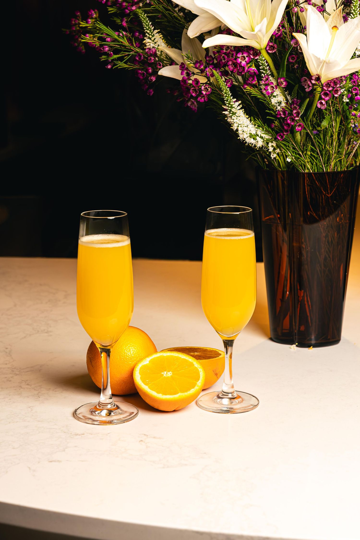 Two champagne flutes filled with orange juice mimosas beside fresh oranges and a floral arrangement on a white surface.