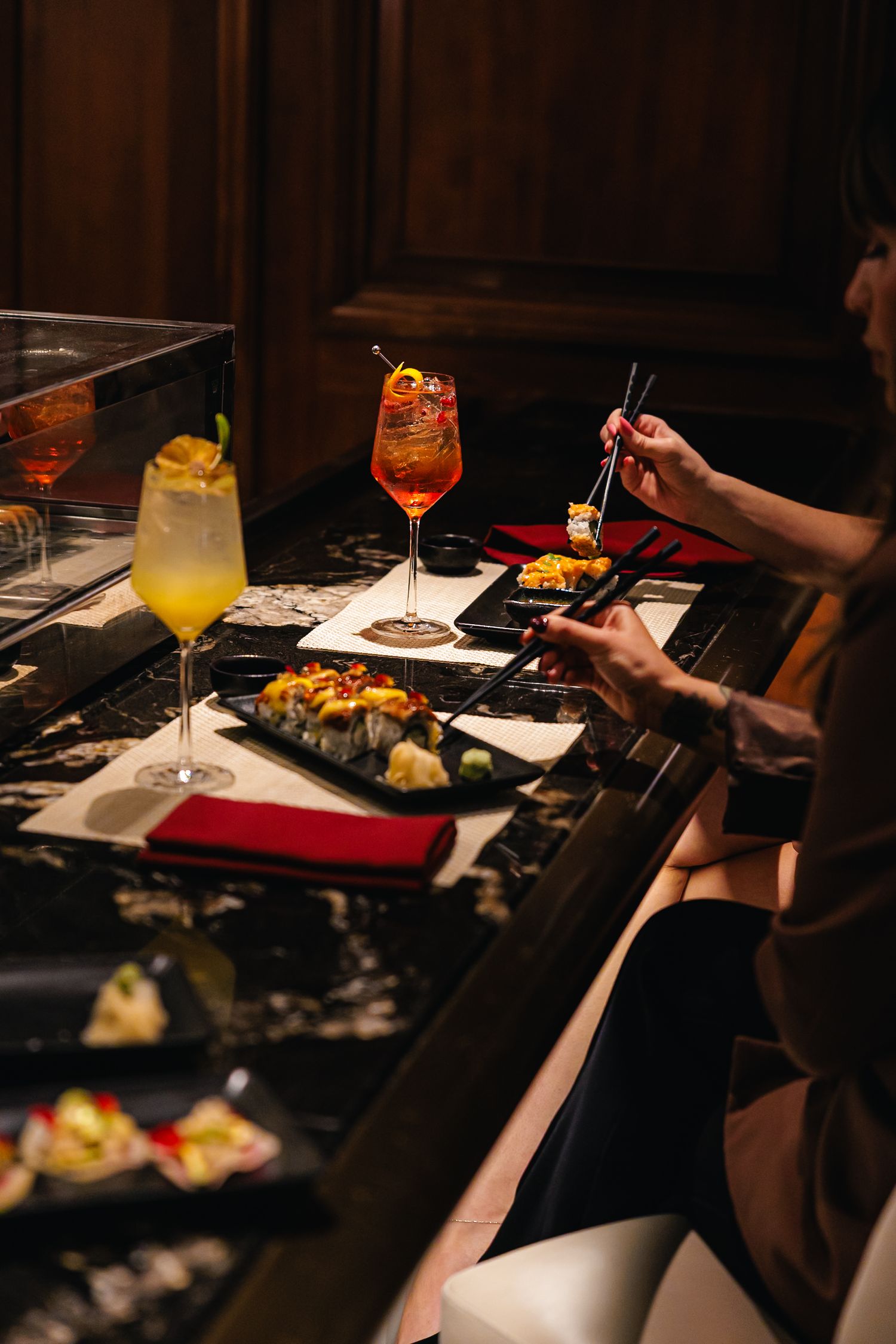 Person using chopsticks to enjoy sushi rolls at a dark marble bar with colorful cocktails including an Aperol Spritz.