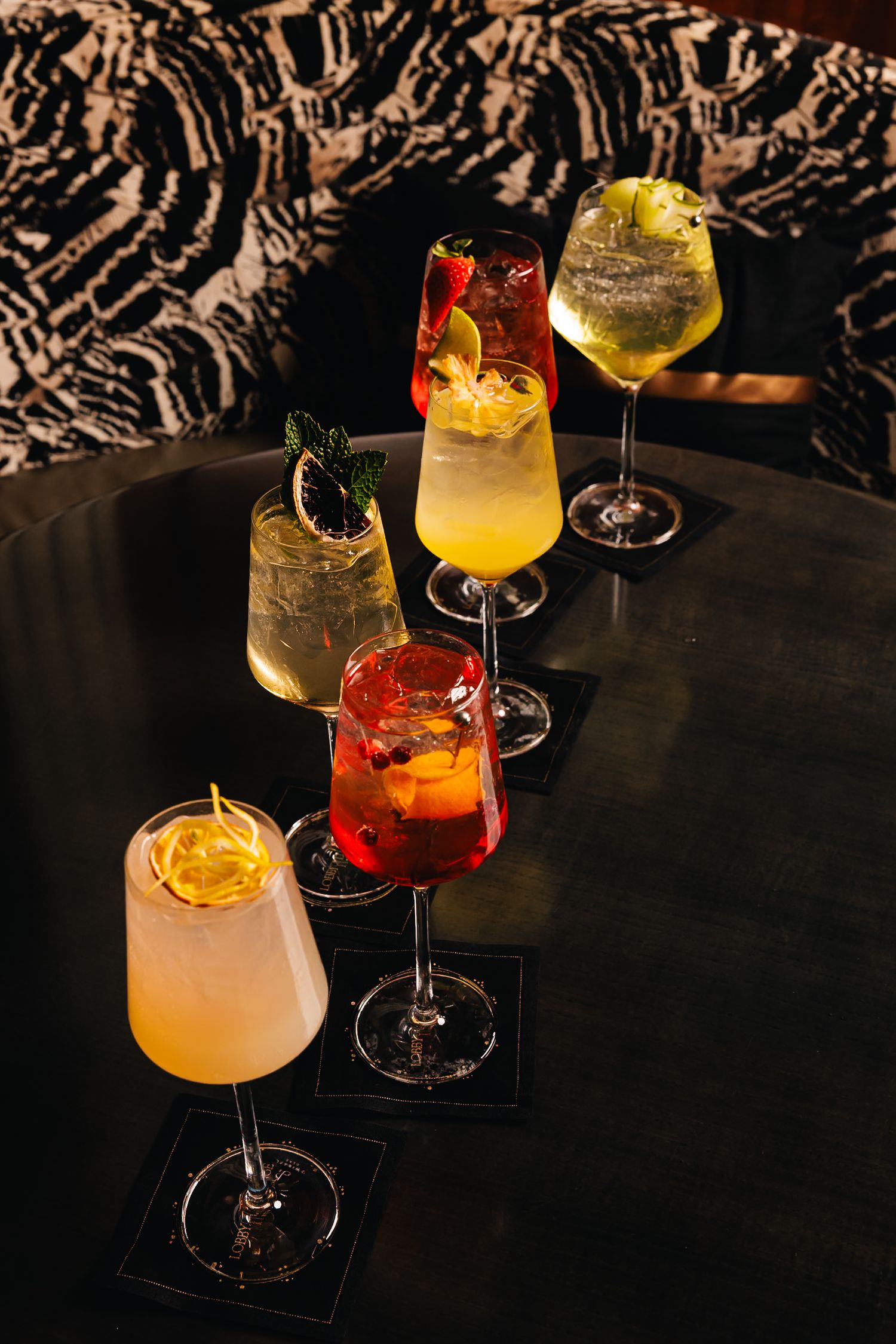 Assorted colorful cocktails in elegant glasses arranged on a dark round table in a stylish bar setting.