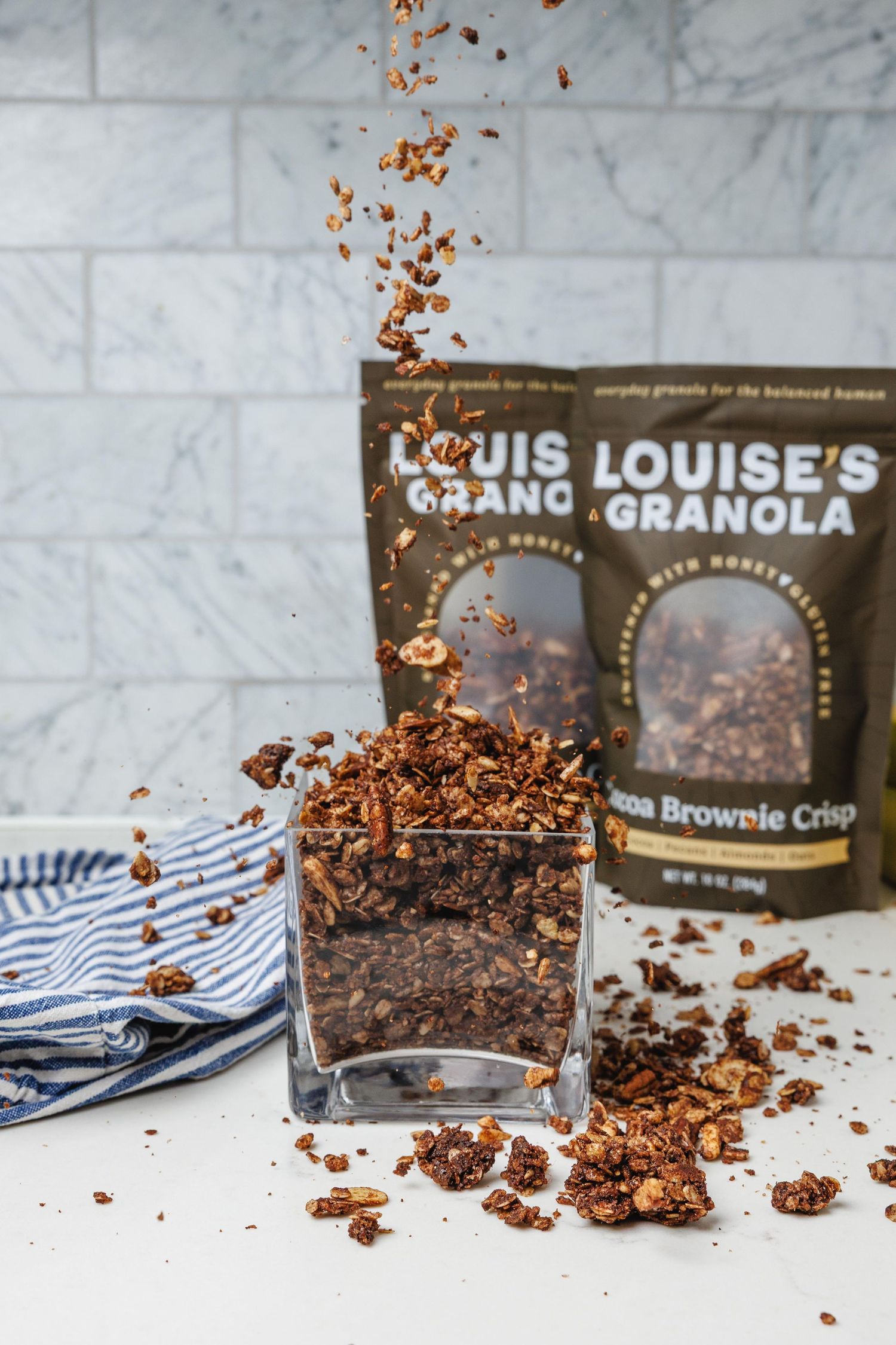 Louise's Granola Cocoa Brownie Crisp pouring into a glass bowl with two dark packaging bags behind it.
