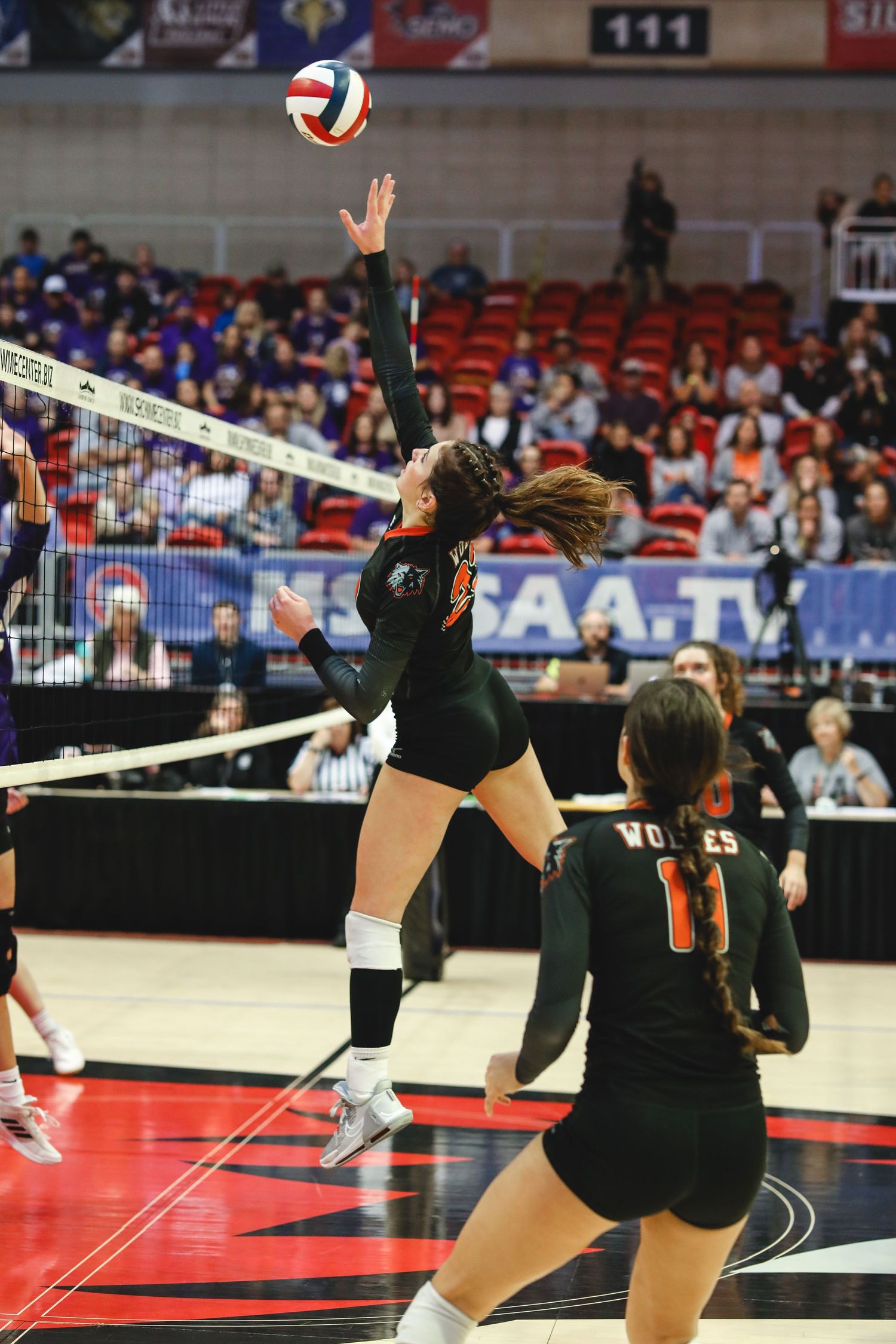 Volleyball players in black uniforms diving and jumping to reach the ball during an intense match.