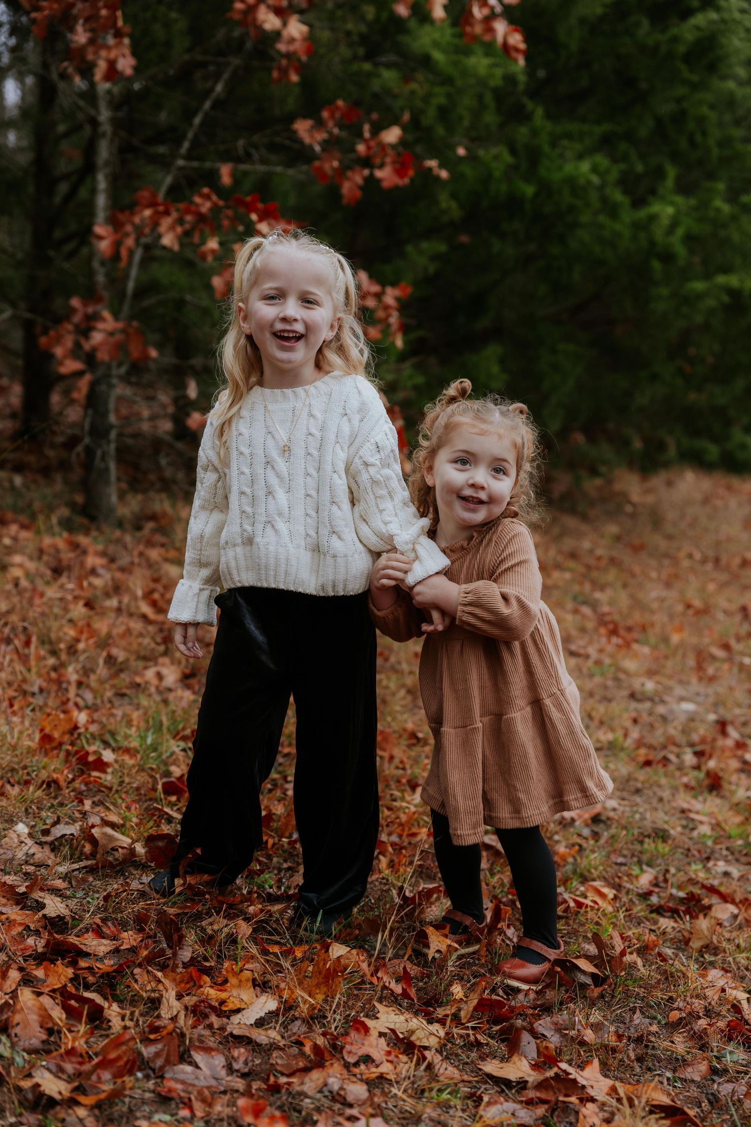 Two children laughing together while standing in autumn leaves wearing cozy sweaters and fall attire.