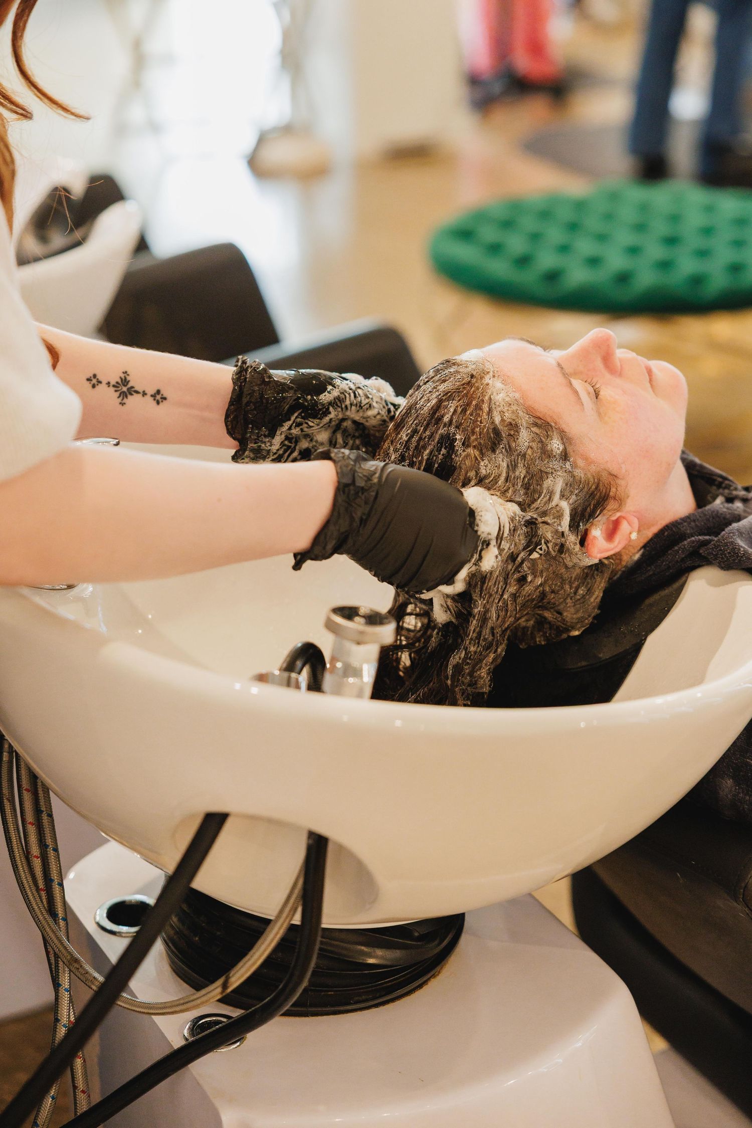 Hair stylist in black gloves washing client hair at salon shampoo bowl, applying lather during professional hair treatment.