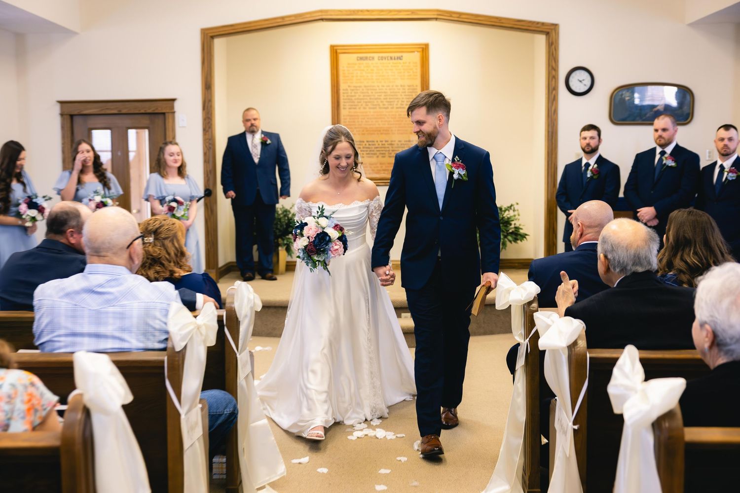 Newlywed couple walks down aisle hand-in-hand past seated guests in a small chapel after their wedding ceremony.
