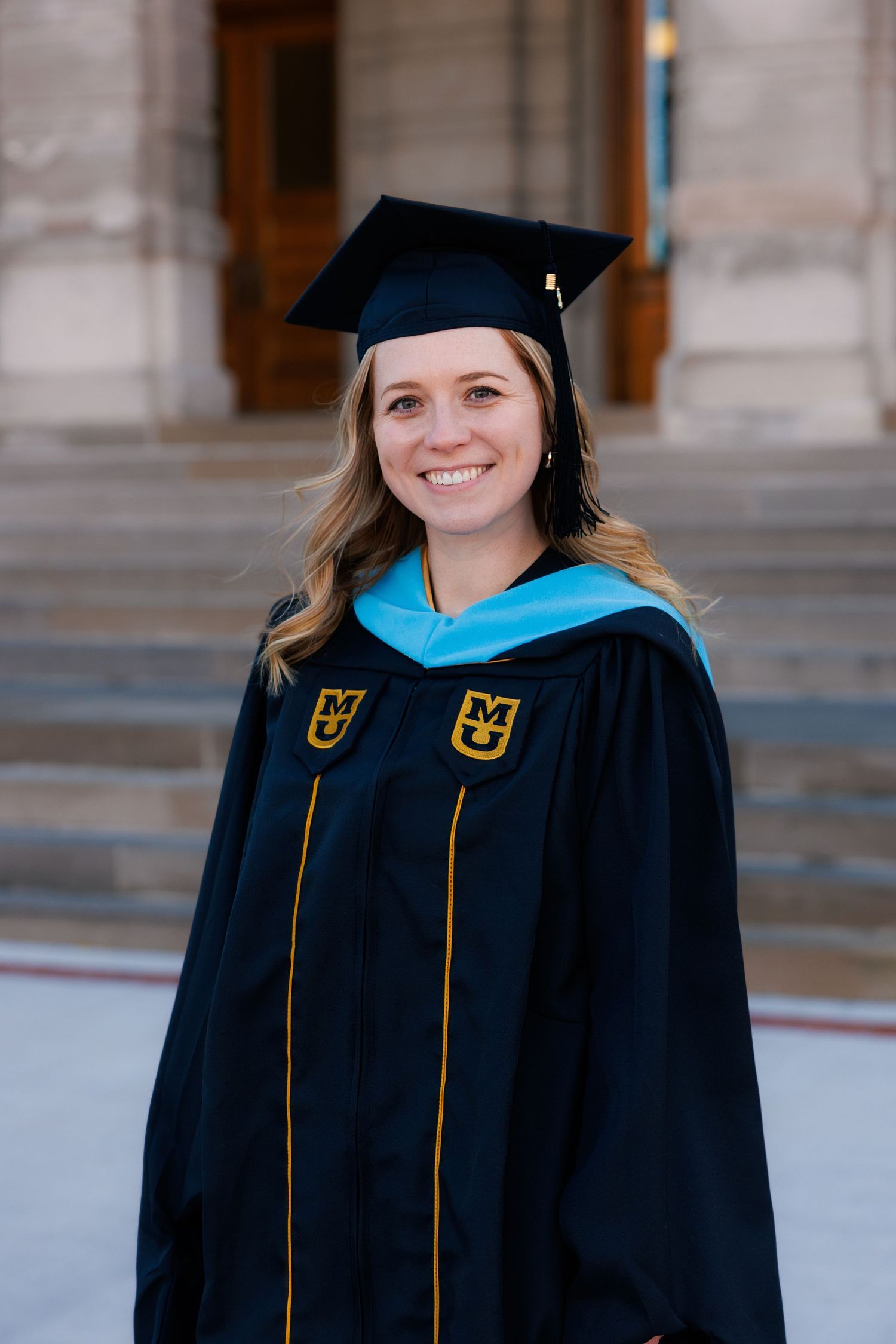 A graduation photo showing someone in black academic regalia with gold trim against stone architecture.