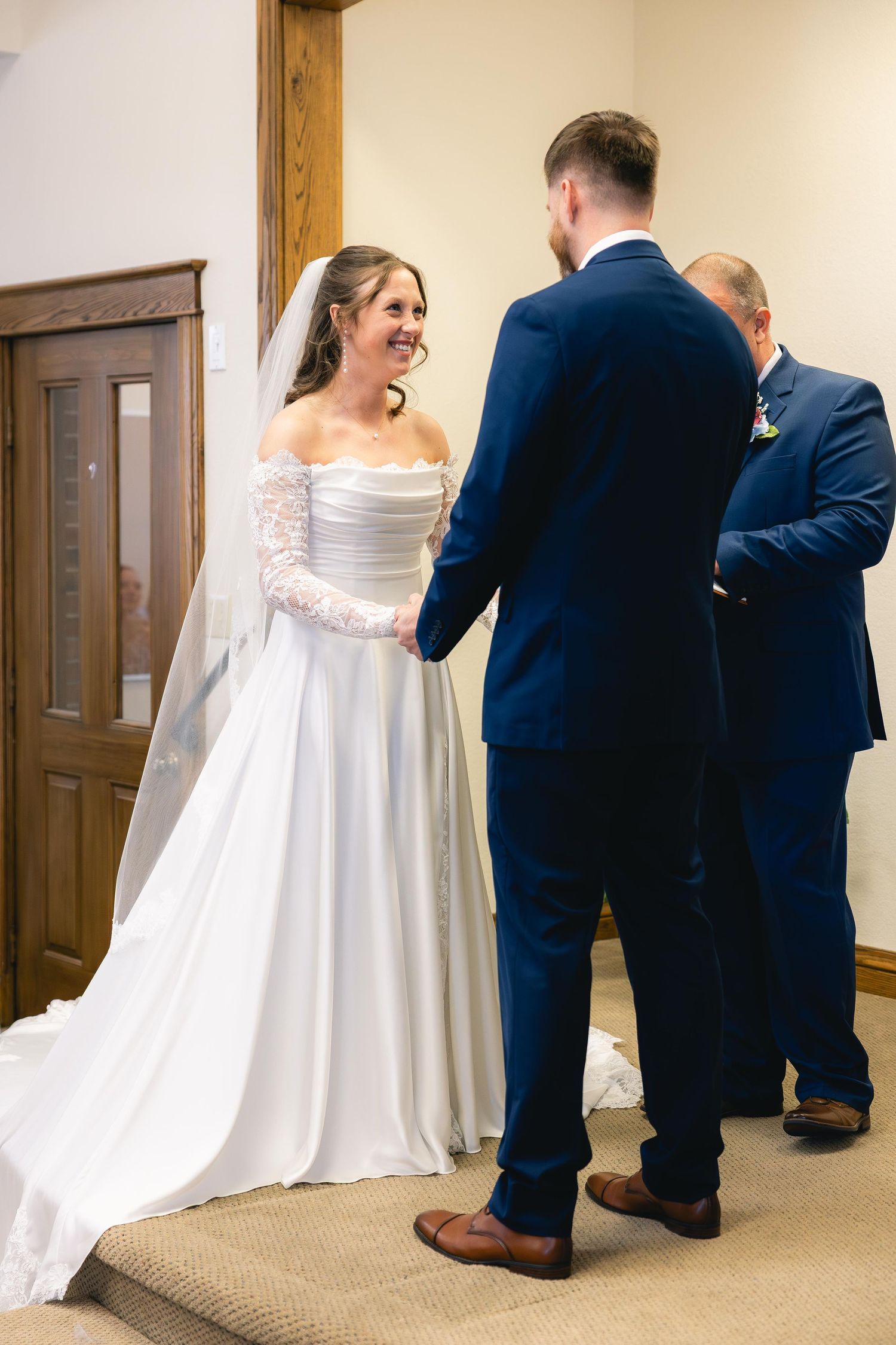 Smiling bride in white off-shoulder gown holds hands with groom in navy suit during indoor wedding ceremony.