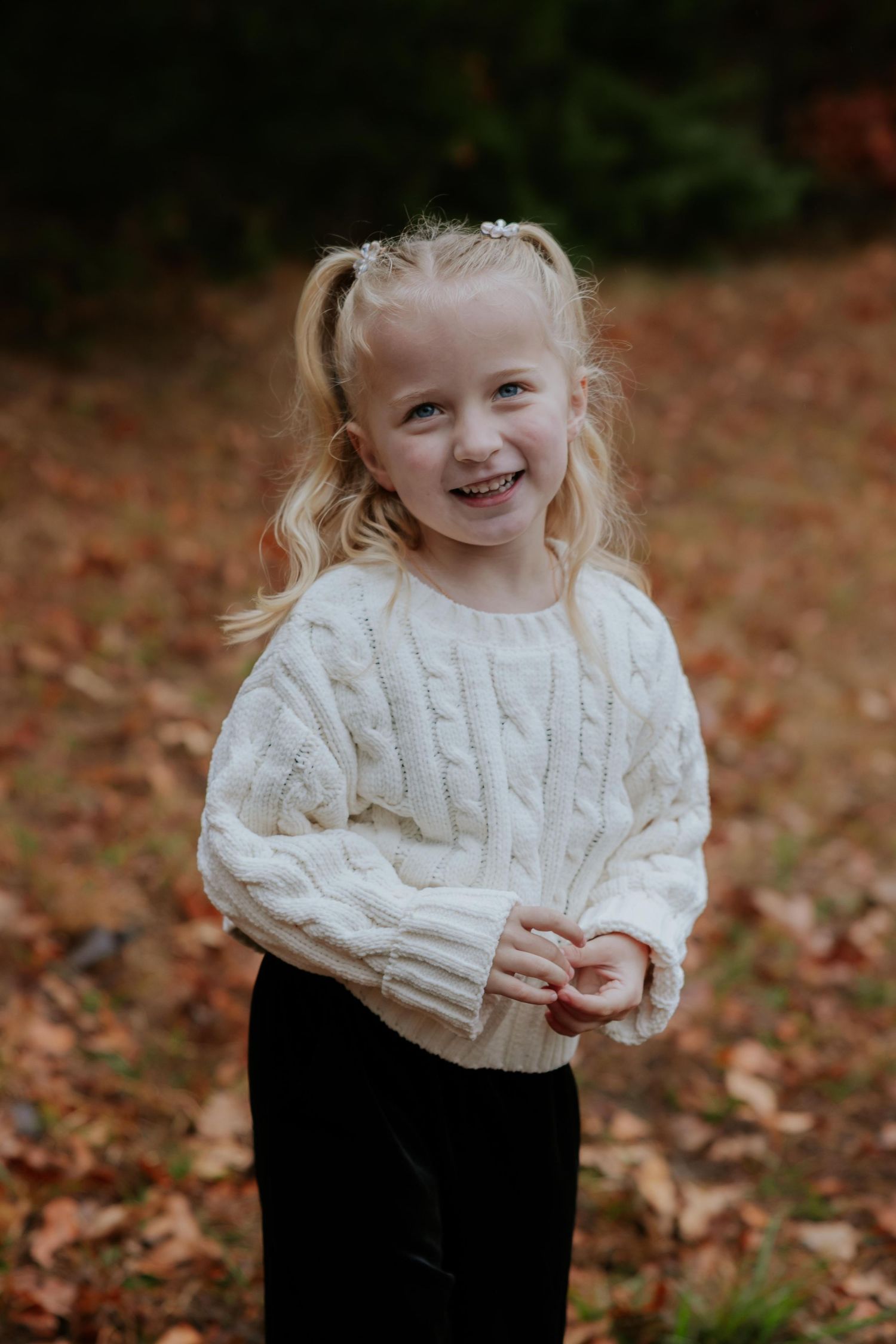 A child in a white cable knit sweater smiles during an outdoor autumn photoshoot.