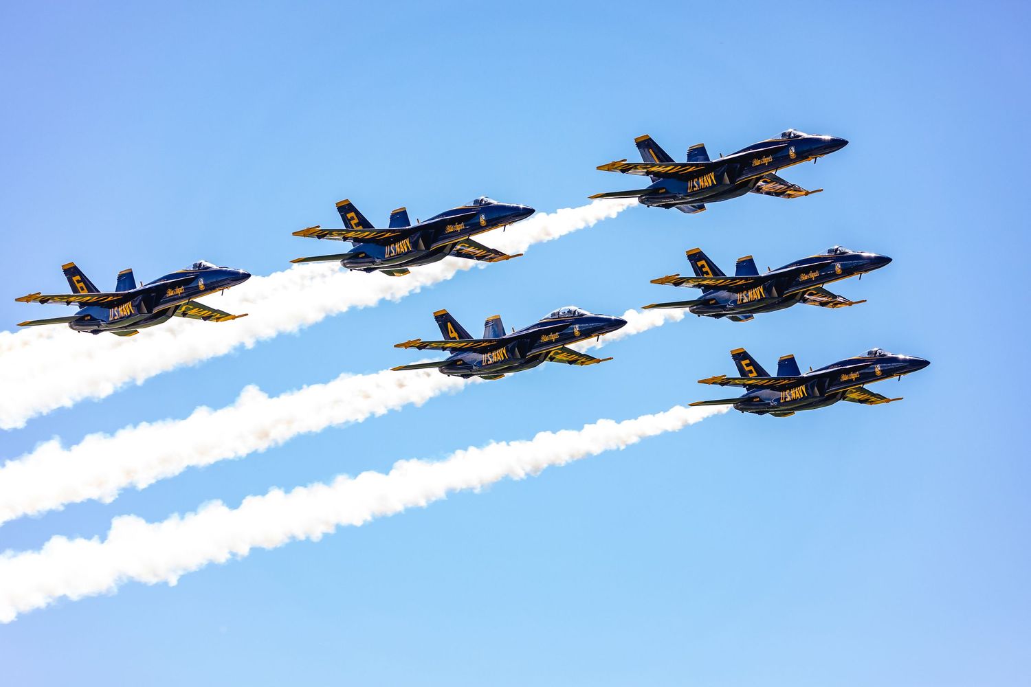 Six Blue Angels jets flying in formation with white contrails against bright blue sky.