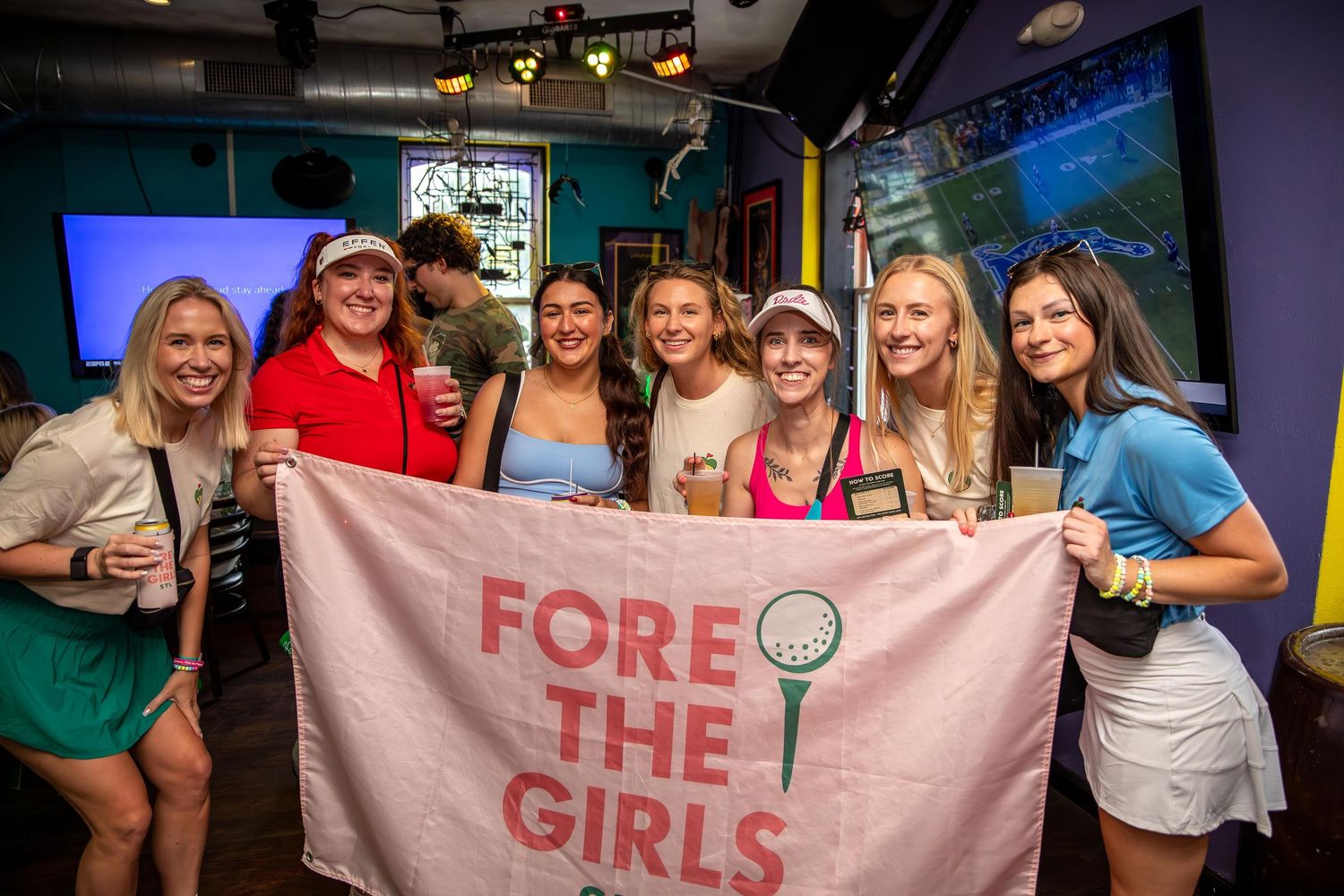 Group of golfers at an event with pink banner in a dimly lit venue.