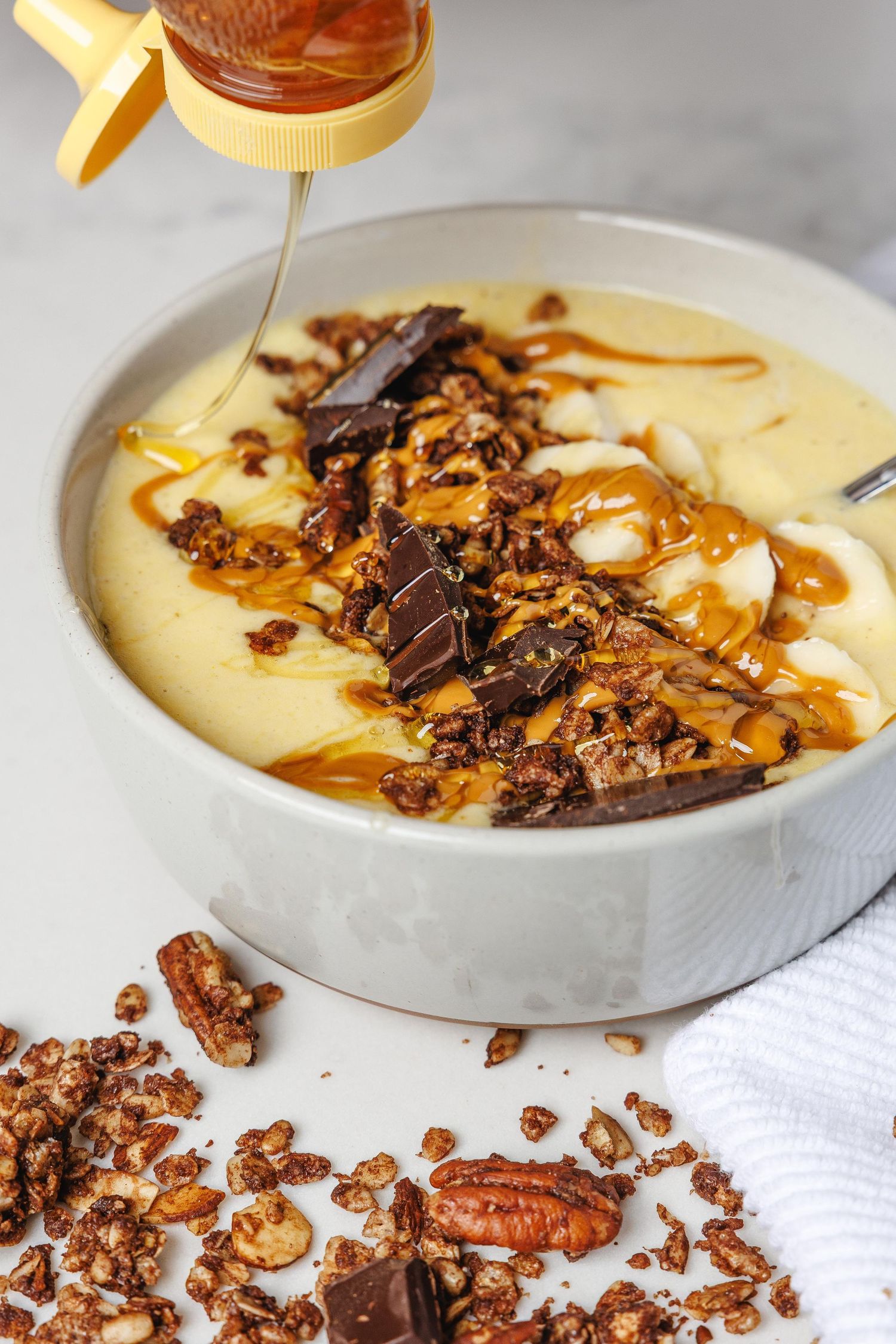 Honey drizzled smoothie bowl topped with granola, dark chocolate chunks, banana slices, and pecans in a gray bowl.