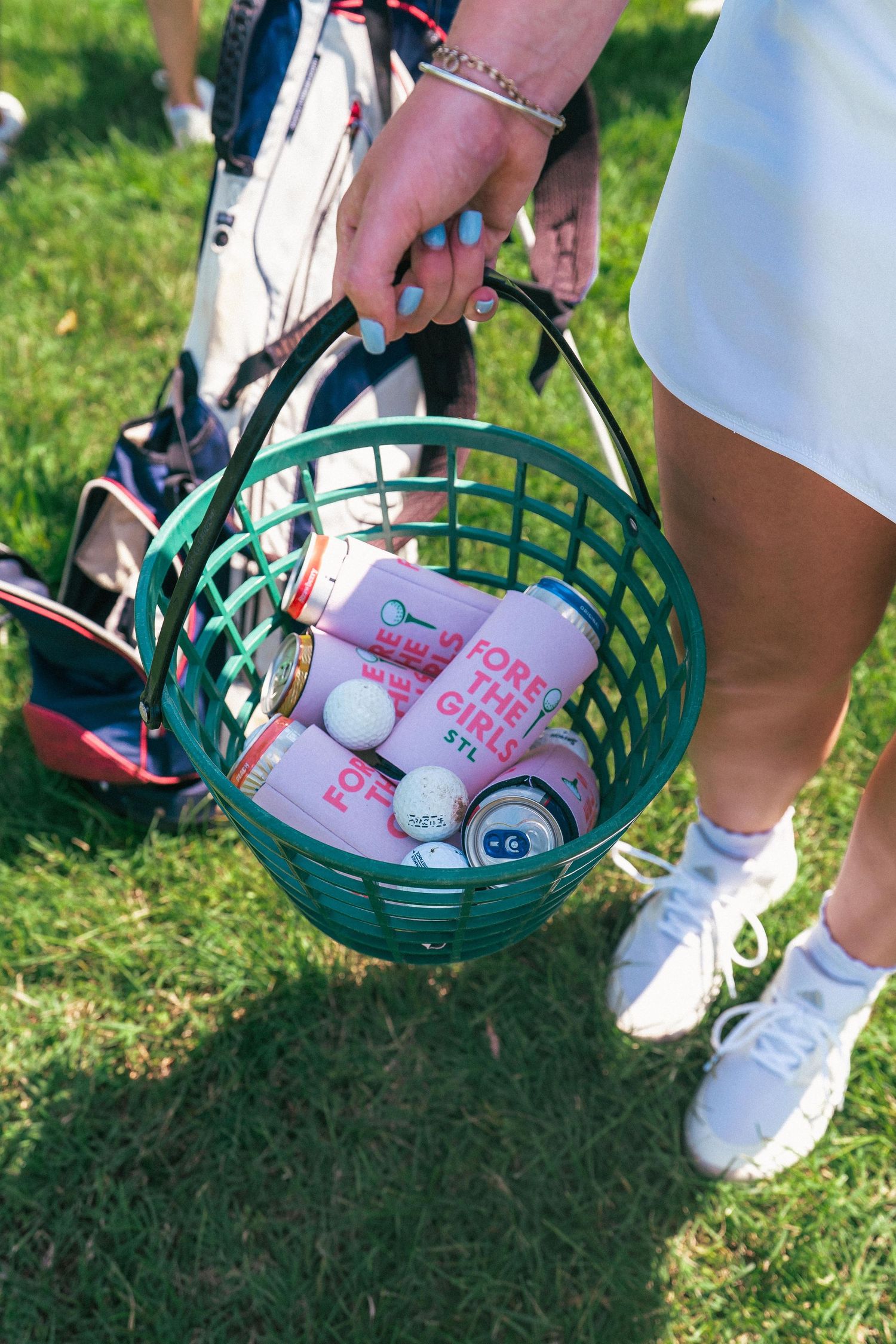 A green basket filled with golf balls and beverages sits on grass next to white sneakers.
